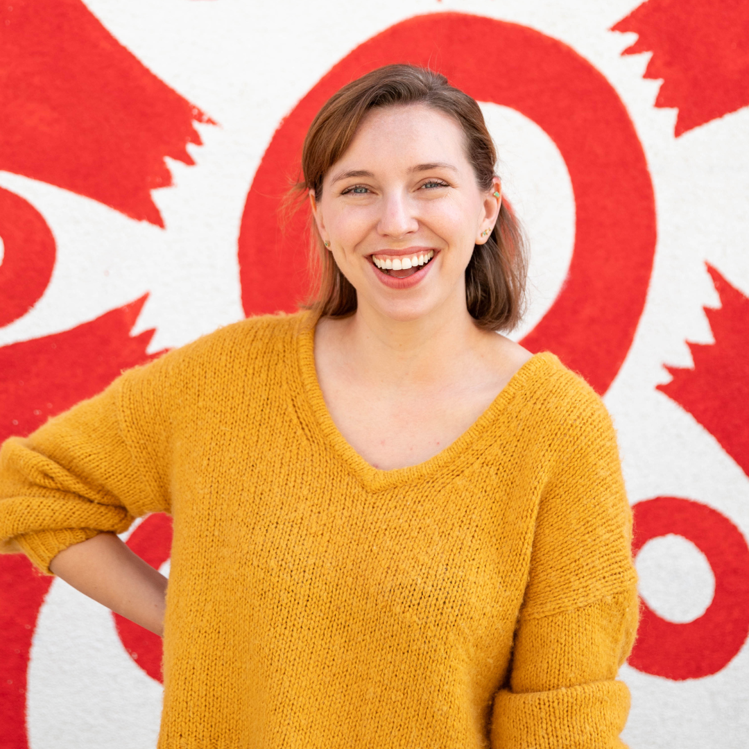 A young woman with short brown hair is smiling and standing in front of a wall painted with red and white abstract designs.
