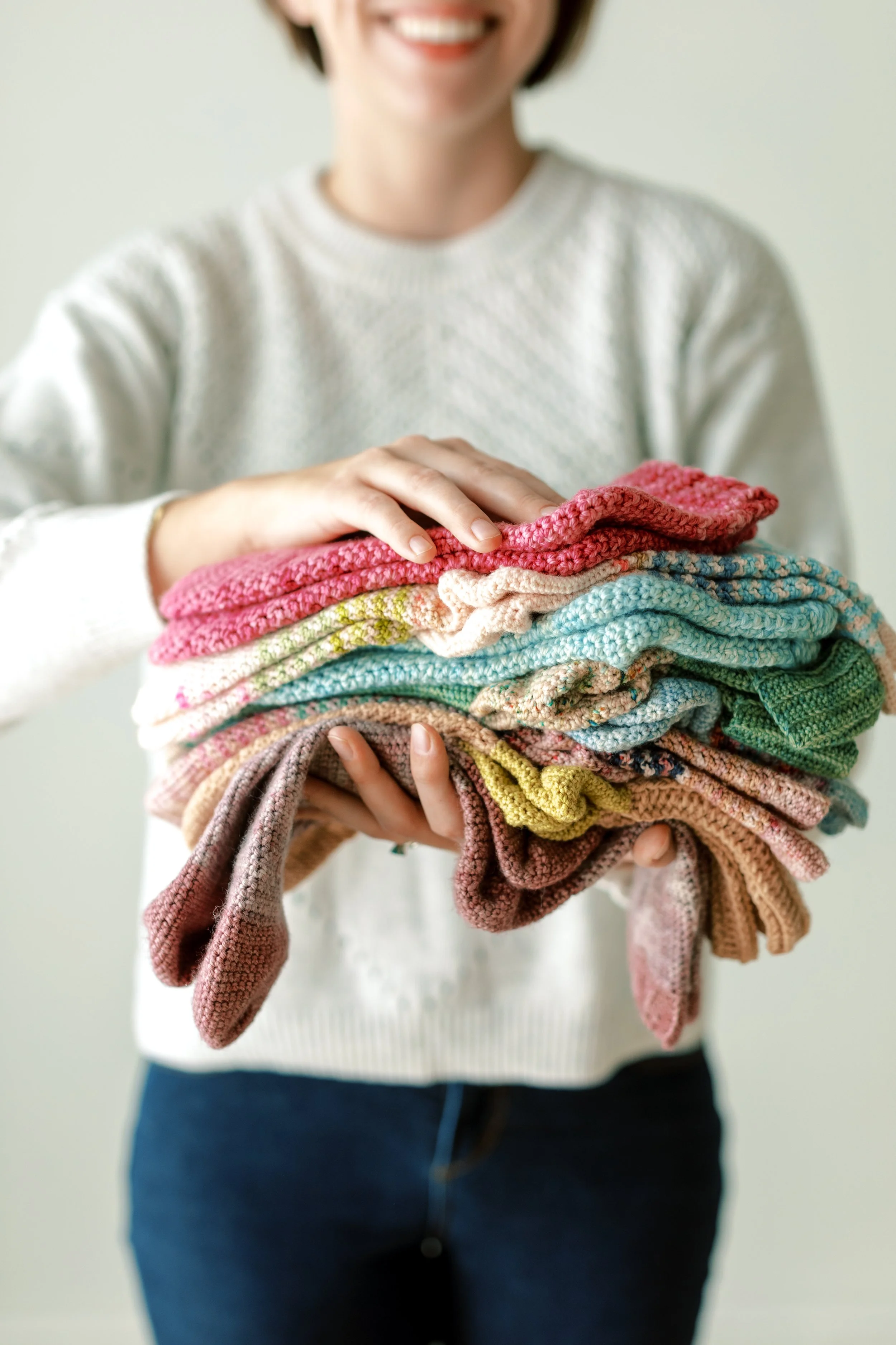 A person holding a large stack of folded colorful sweaters, smiling.