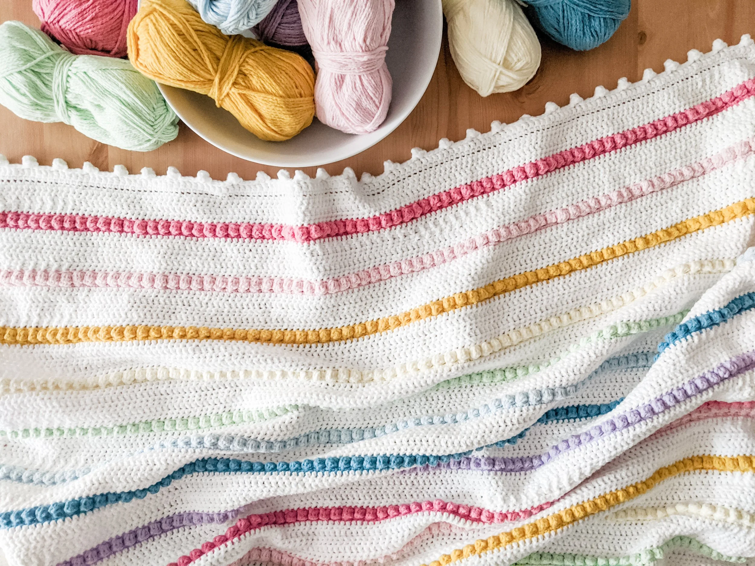 Colorful yarn balls in a bowl and a neatly folded multicolored crocheted blanket with various pastel and bright colors on a wooden surface.