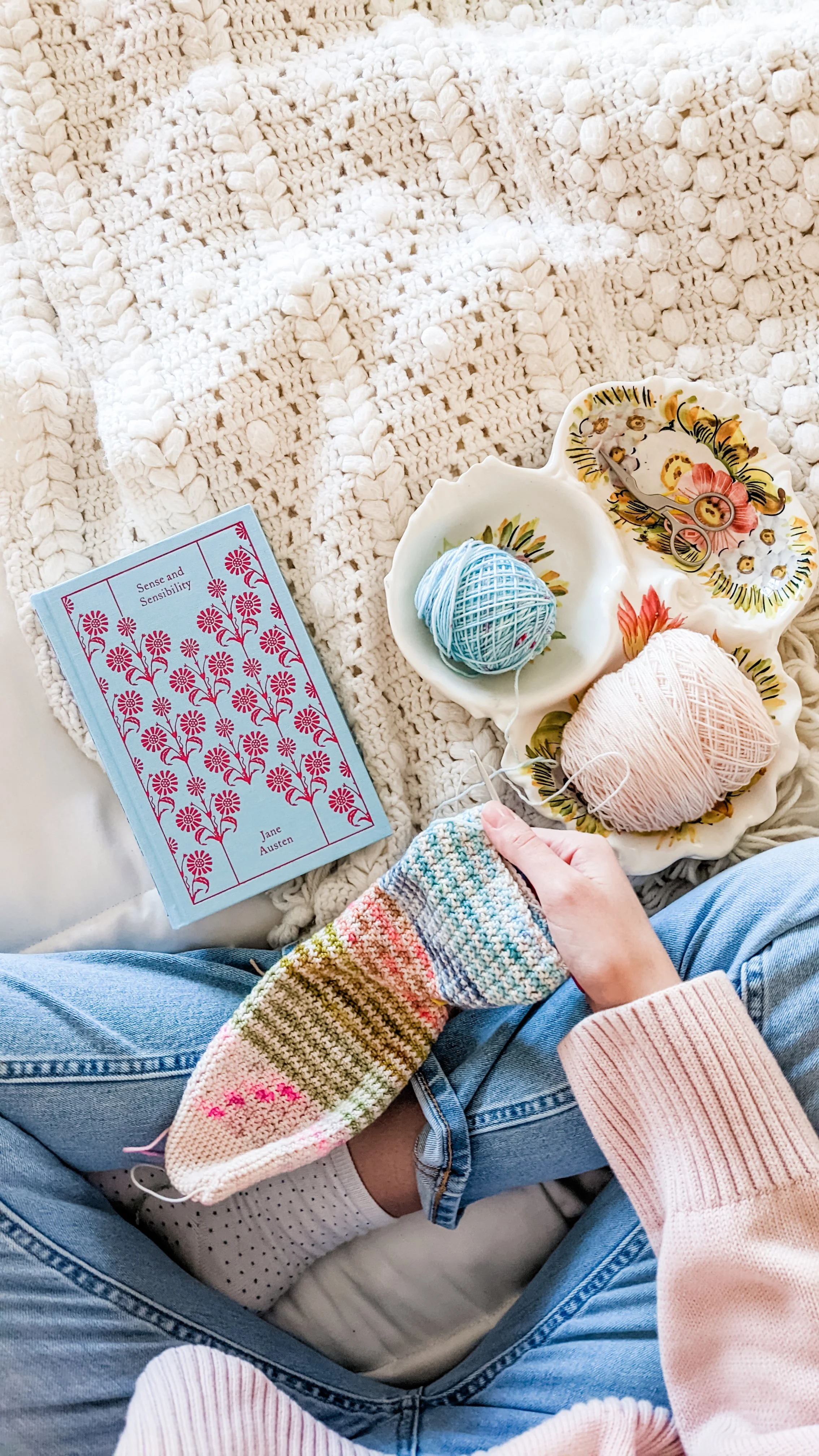 Person sitting on a bed with a cream-colored chunky knit blanket, holding colorful striped socks and knitting supplies. On the bed, there's a plate with yarn and scissors, a book titled 'Sense and Sensibility' by Jane Austen, and additional yarn nearby.