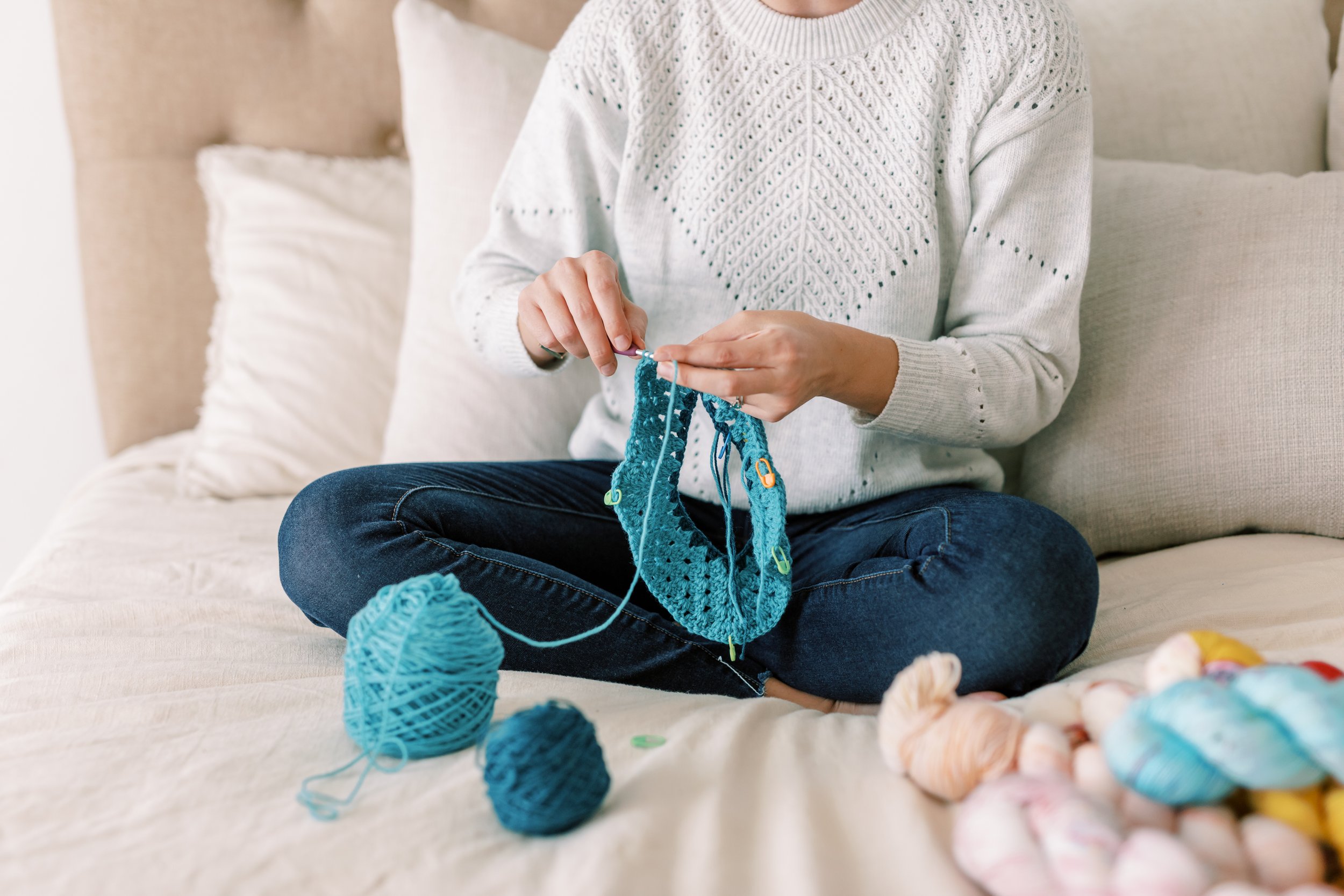 Person knitting a blue garment while sitting on a beige couch with yarn and knitting supplies around them.