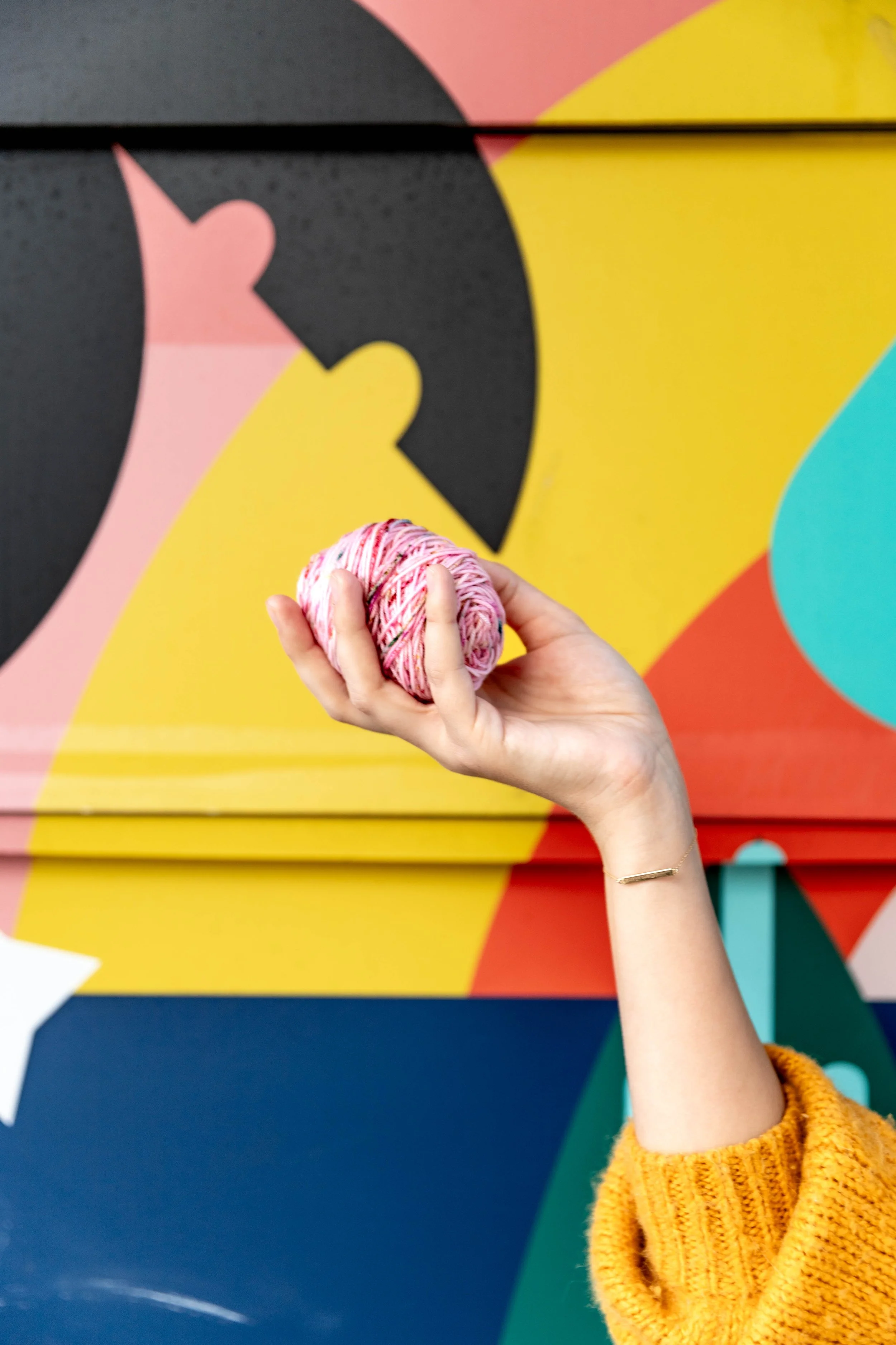Close-up of a person holding a pink and white yarn ball with a yellow, pink, black, and blue abstract mural background.