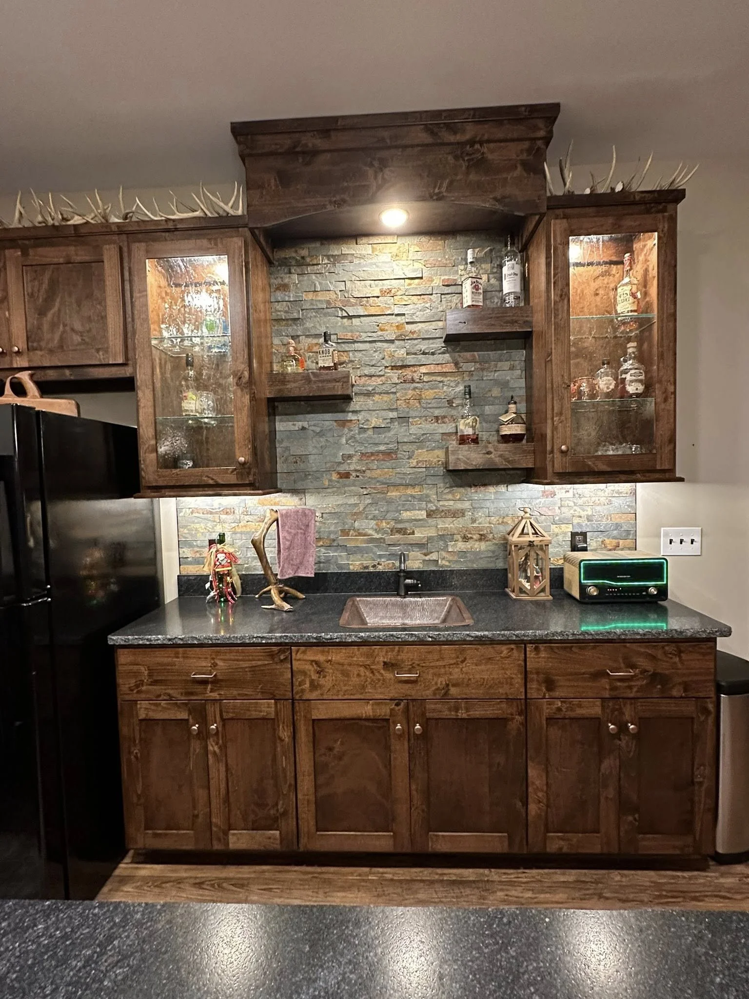 Rustic kitchen with dark granite countertops, wooden cabinets, and a brick backsplash. Open shelves hold liquor bottles, and antlers are displayed on top of the cabinets. A small sink is in the center of the counter, with various decorations and a ra
