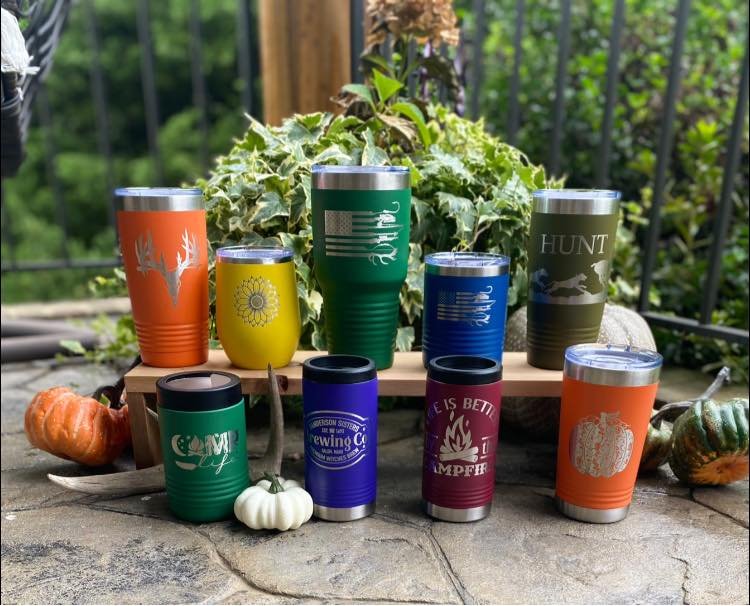 Collection of colorful insulated drink tumblers with various designs and sayings, displayed outdoors on a stone surface with pumpkins and greenery in the background.