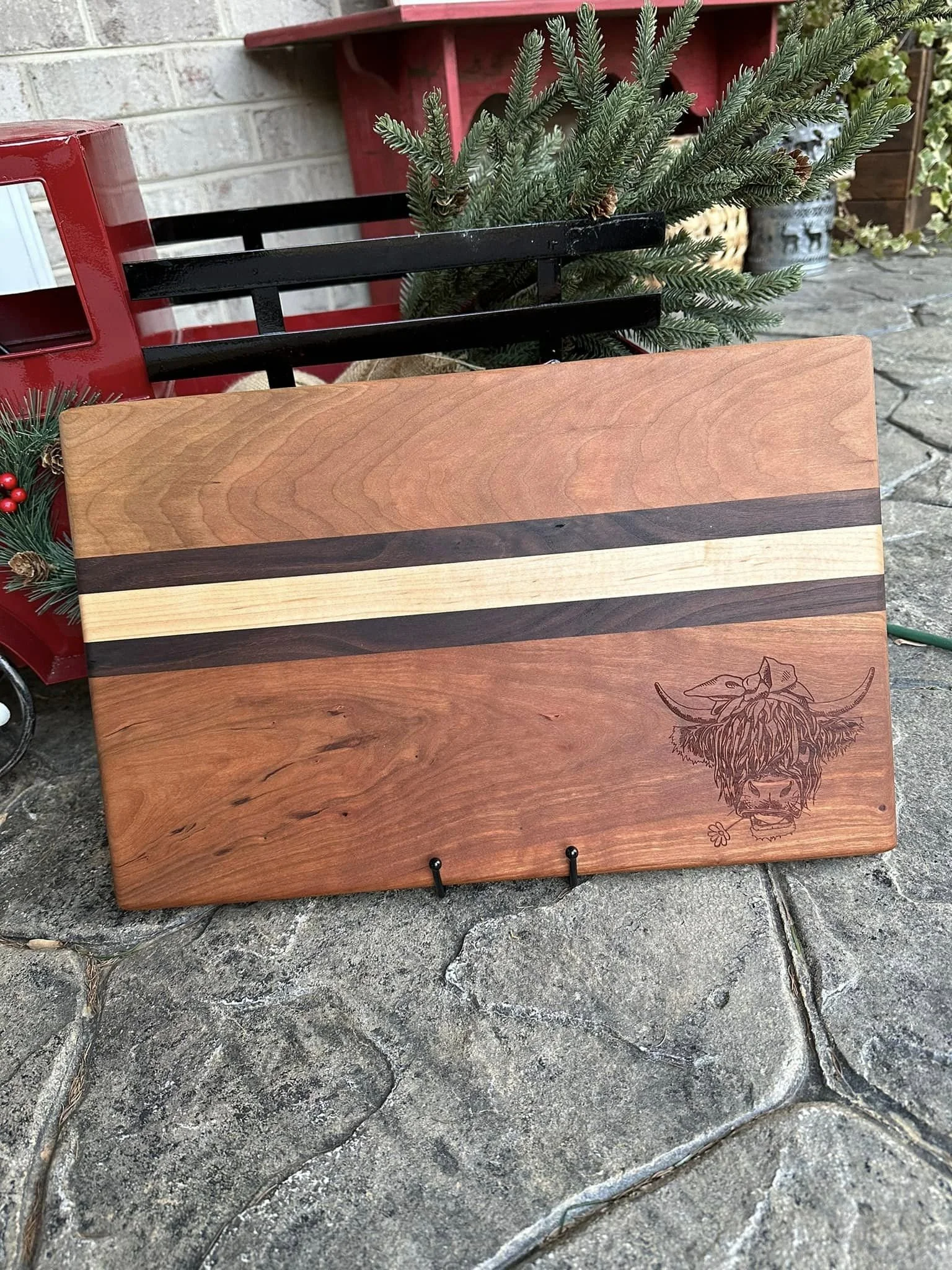 A wooden cutting board with a Highland cow engraved on the bottom right corner, placed outdoors on a stone surface. Behind the board, there is a small pine tree, a red decorative cart with Christmas-themed pinecones and berries, and other outdoor dec
