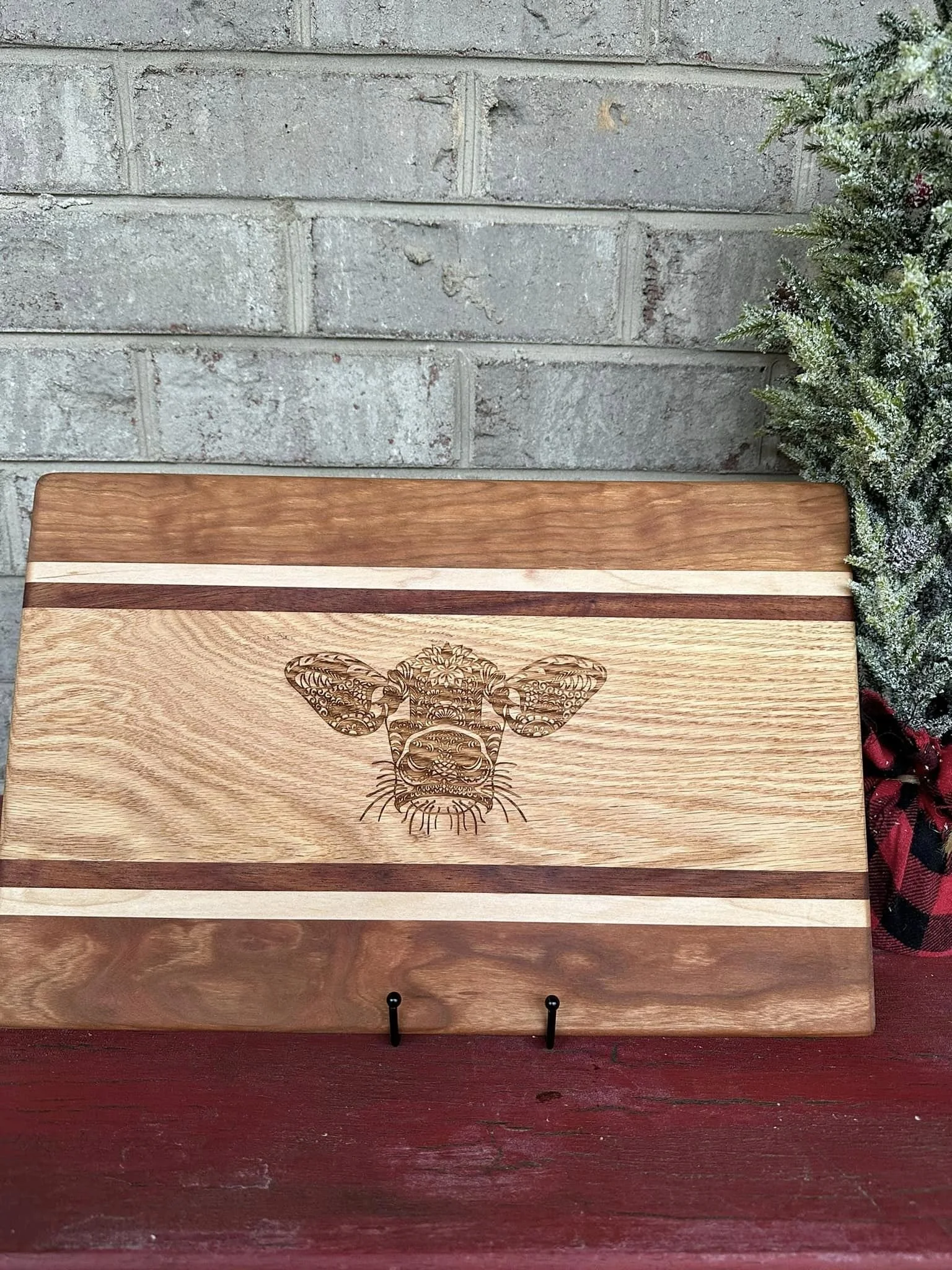 Wooden cutting board with a laser-engraved cow's face and decorative patterns, resting on a red surface against a brick wall, with a Christmas themed decoration partially visible on the right side.
