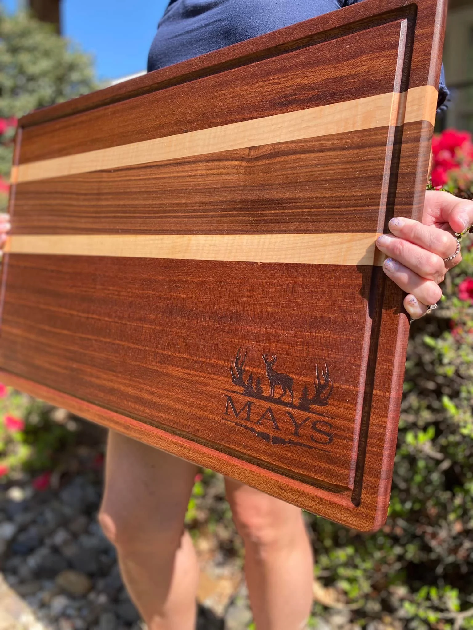 Person holding a wooden cutting board with a striped design and a deer logo with the word "MAYS" engraved on it, outdoors with flowers in the background.