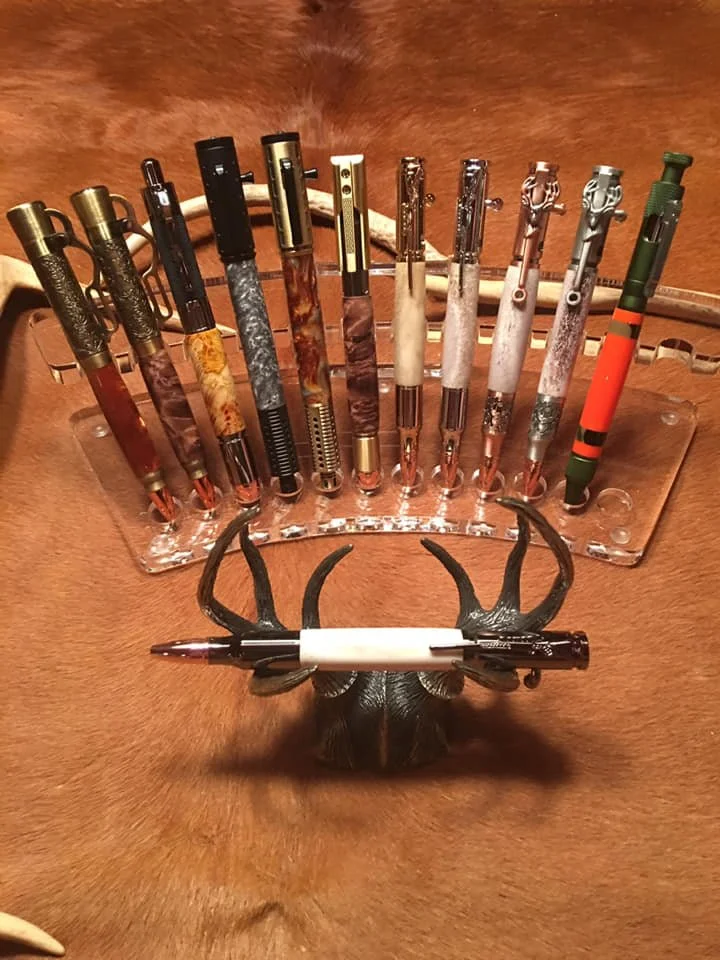 A collection of decorative pens with various colored and textured barrels, displayed in a clear holder on a wooden surface. In front of the pens, there is a decorative item resembling antlers with a black-and-white pen resting on it.