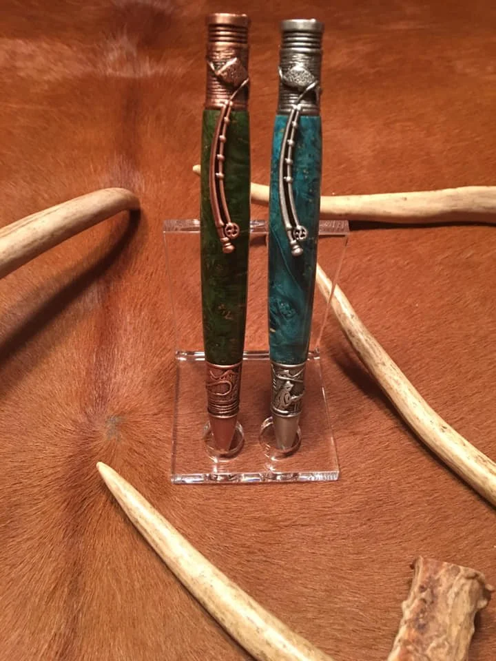 Two decorative knives with turquoise and green handles, metal accents, and shaped handles, displayed on a clear stand on a wooden surface with antlers around.