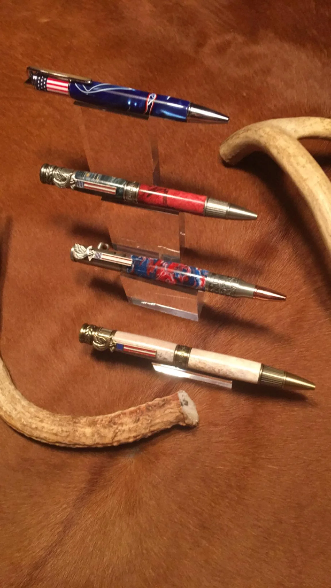 Four decorative pens with patriotic designs, placed on a reddish-brown surface, with two animal horns, one curved with a light color and the other with a darker, rougher texture nearby.