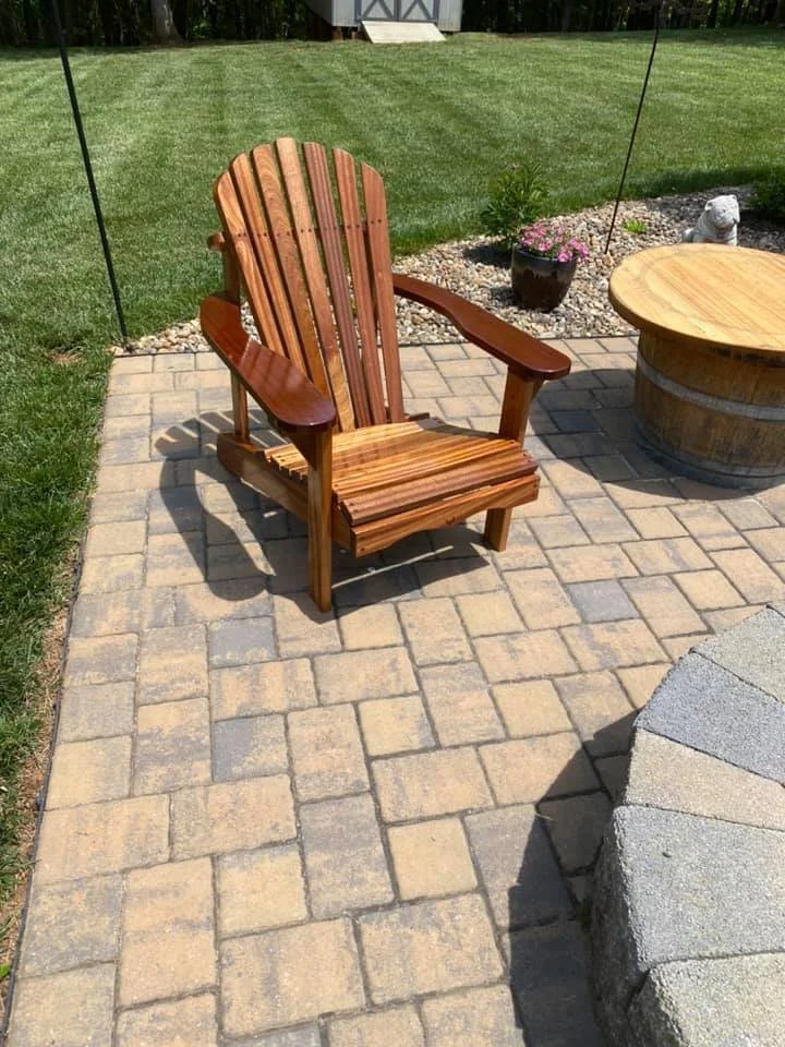 A wooden Adirondack chair on a paved patio area with green grass, a flower pot, and a garden statue in the background.