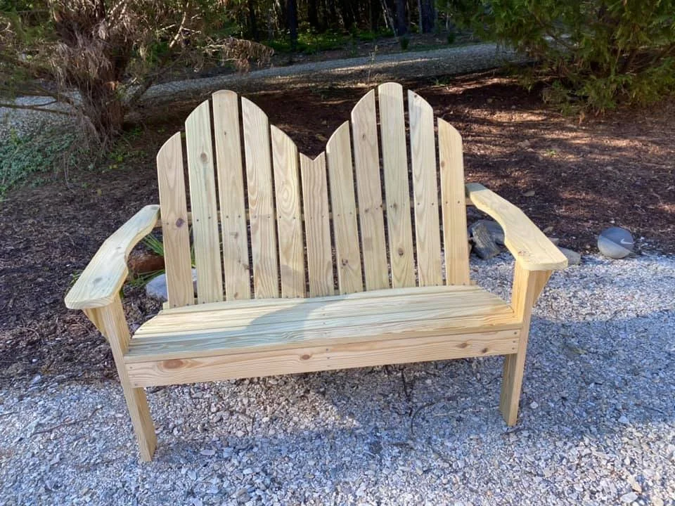 A wooden bench with a high backrest made of vertical slats, placed outdoors on a gravel surface with trees in the background.
