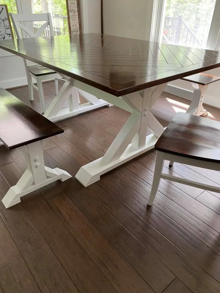 Wood dining table with white painted trestle base, surrounded by matching wooden chairs and benches, in a bright room with hardwood floors and large windows.