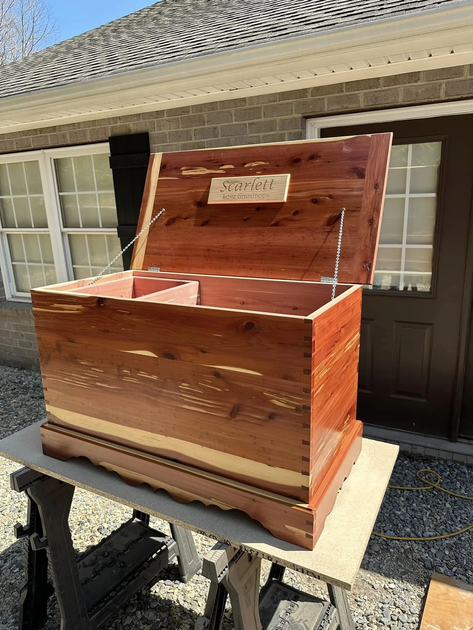 Unfinished wooden memory box with a hinged lid, inscribed with the name 'Scarlett' and the phrase 'Love Grandpops', placed on a workbench outside a brick house.