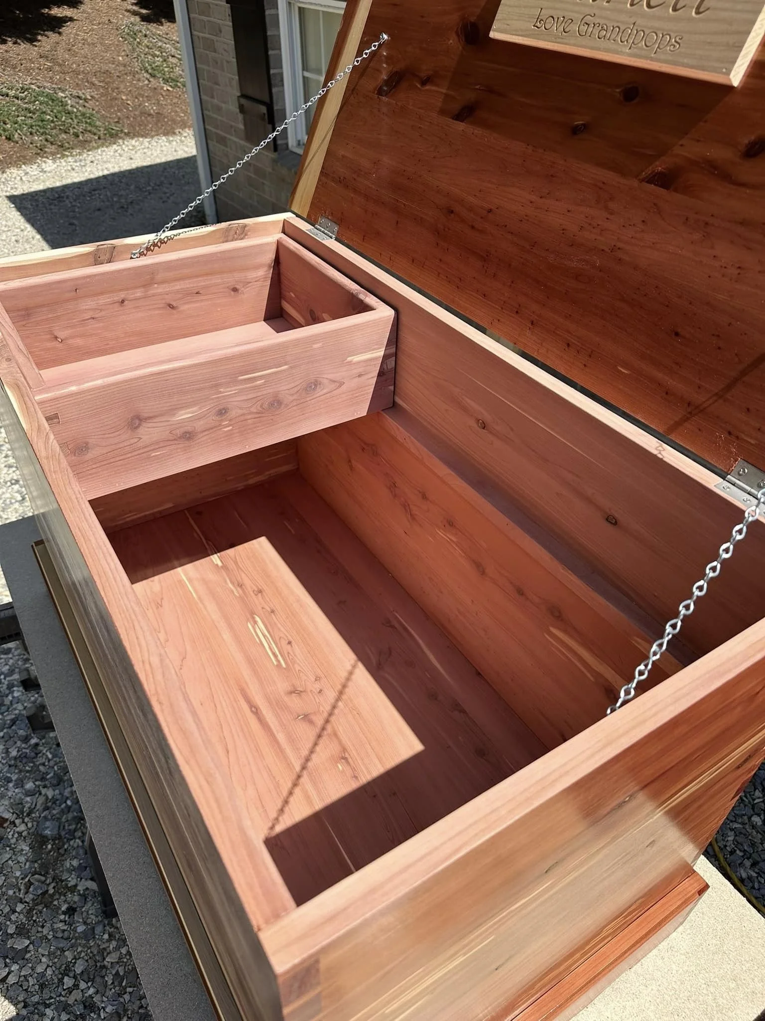 An open wooden treasure chest with a smaller compartment inside, outdoors on a gravel surface near a brick building.