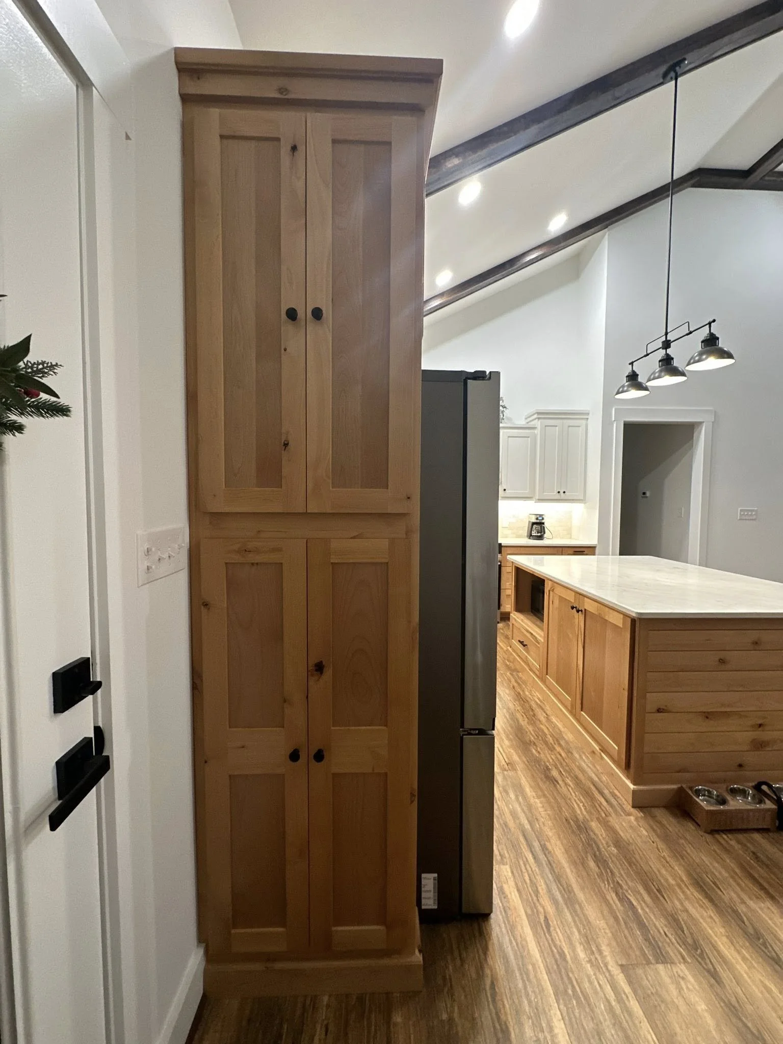 A tall wooden cabinet with double doors and black knobs, situated next to a stainless steel refrigerator in a kitchen with wooden flooring and white cabinetry.