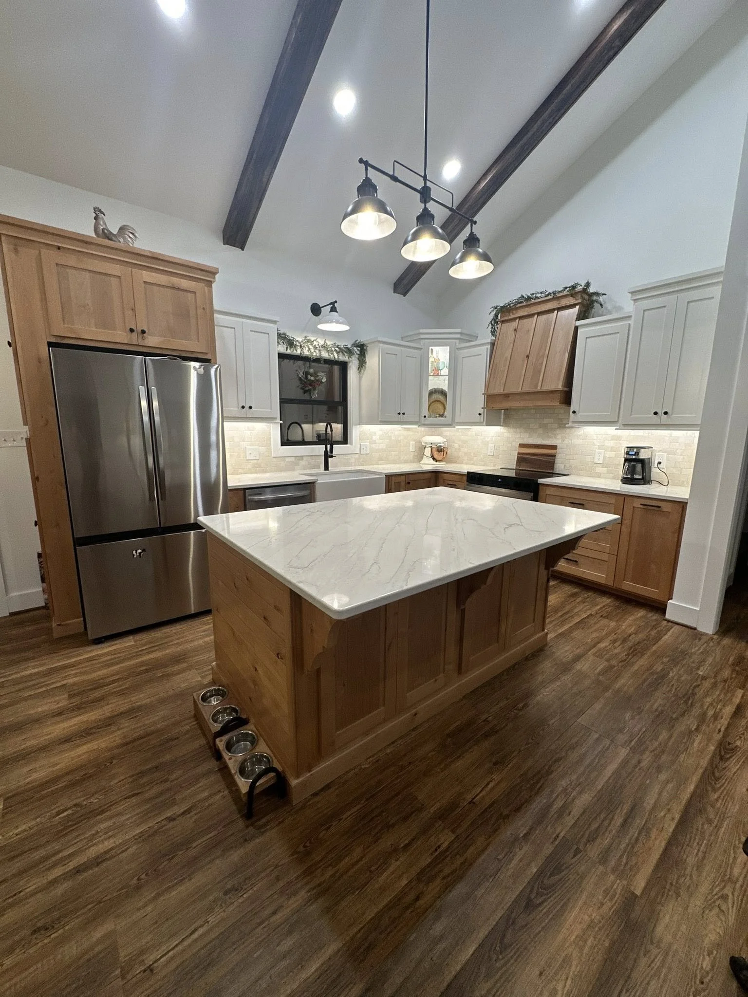 A modern kitchen with white and wood cabinetry, a large marble island, stainless steel fridge, wood flooring, and high vaulted ceiling with exposed dark beams and hanging industrial-style lights.