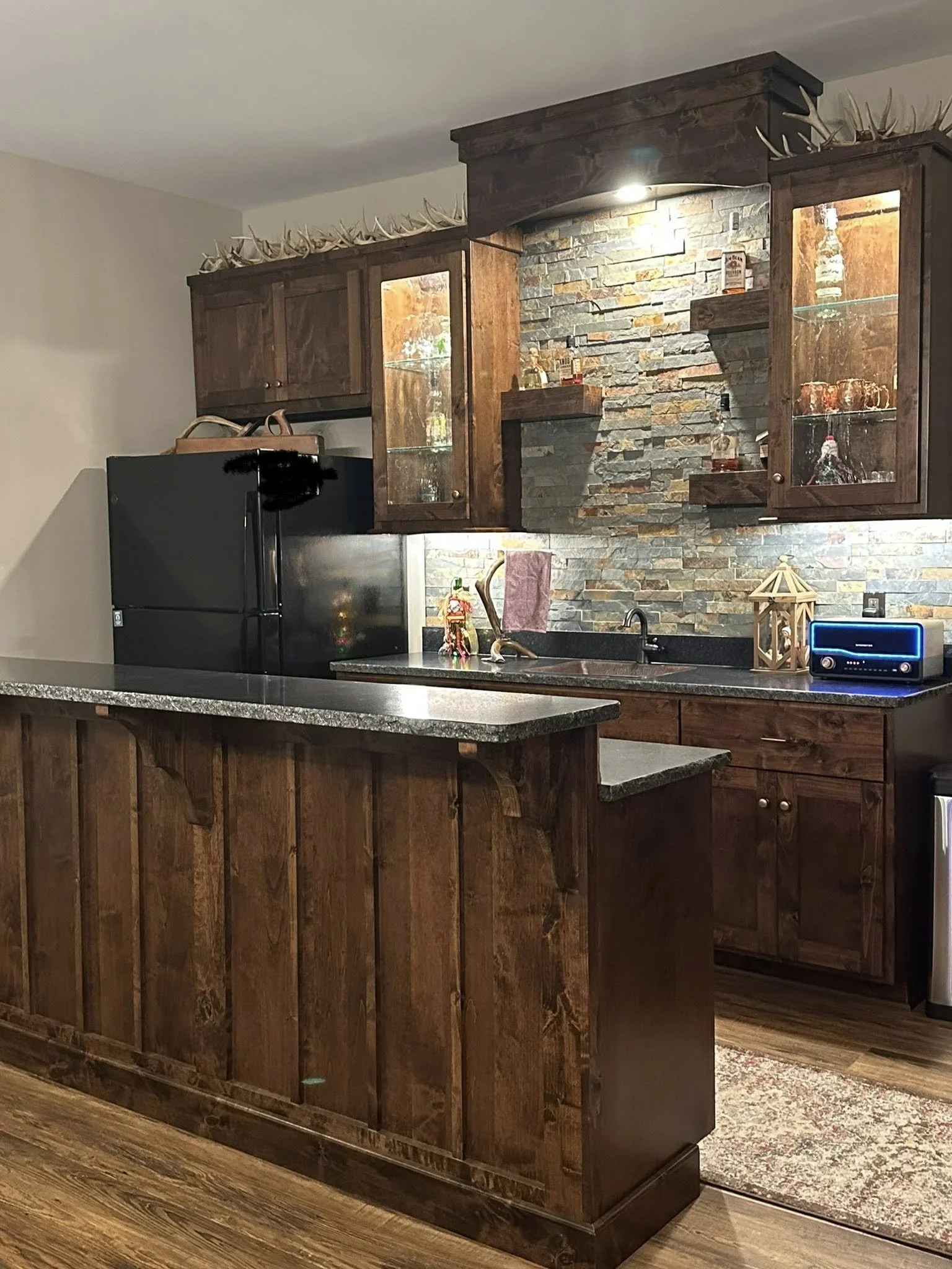 Rustic kitchen with wooden cabinets, a black refrigerator, a granite countertop, and a brick backsplash. Decorative items include a nativity scene, a miniature house, and a small blue radio.