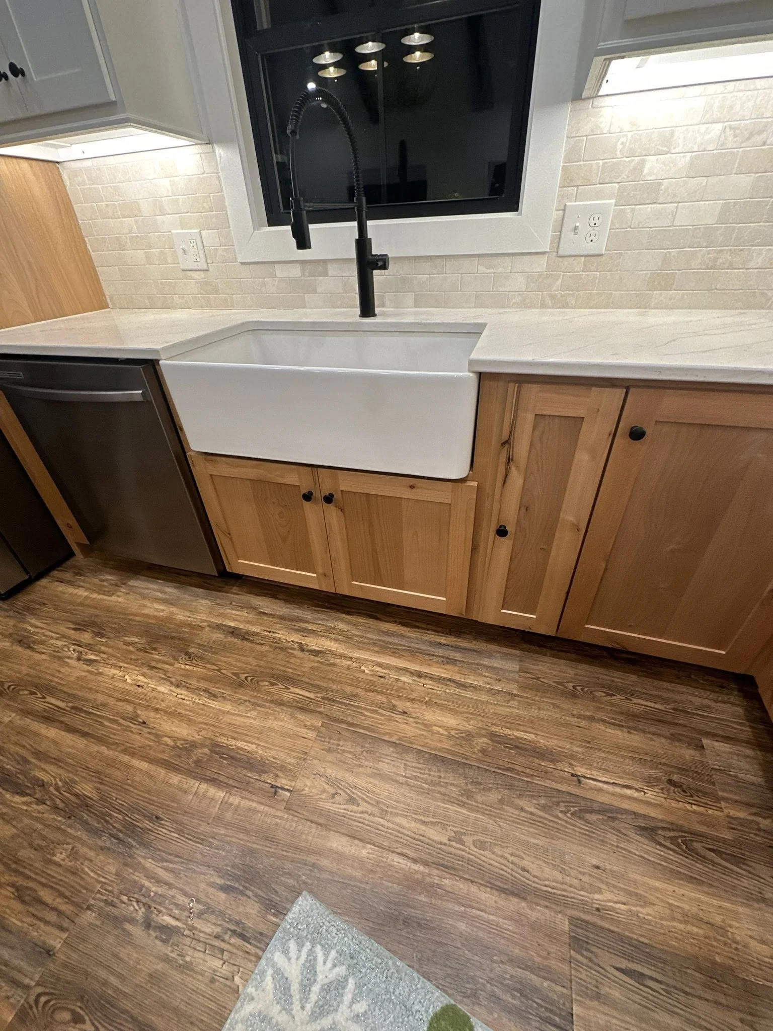 Kitchen sink with black gooseneck faucet, wooden cabinets, light-colored countertop, beige brick backsplash, window above the sink, and hardwood flooring.