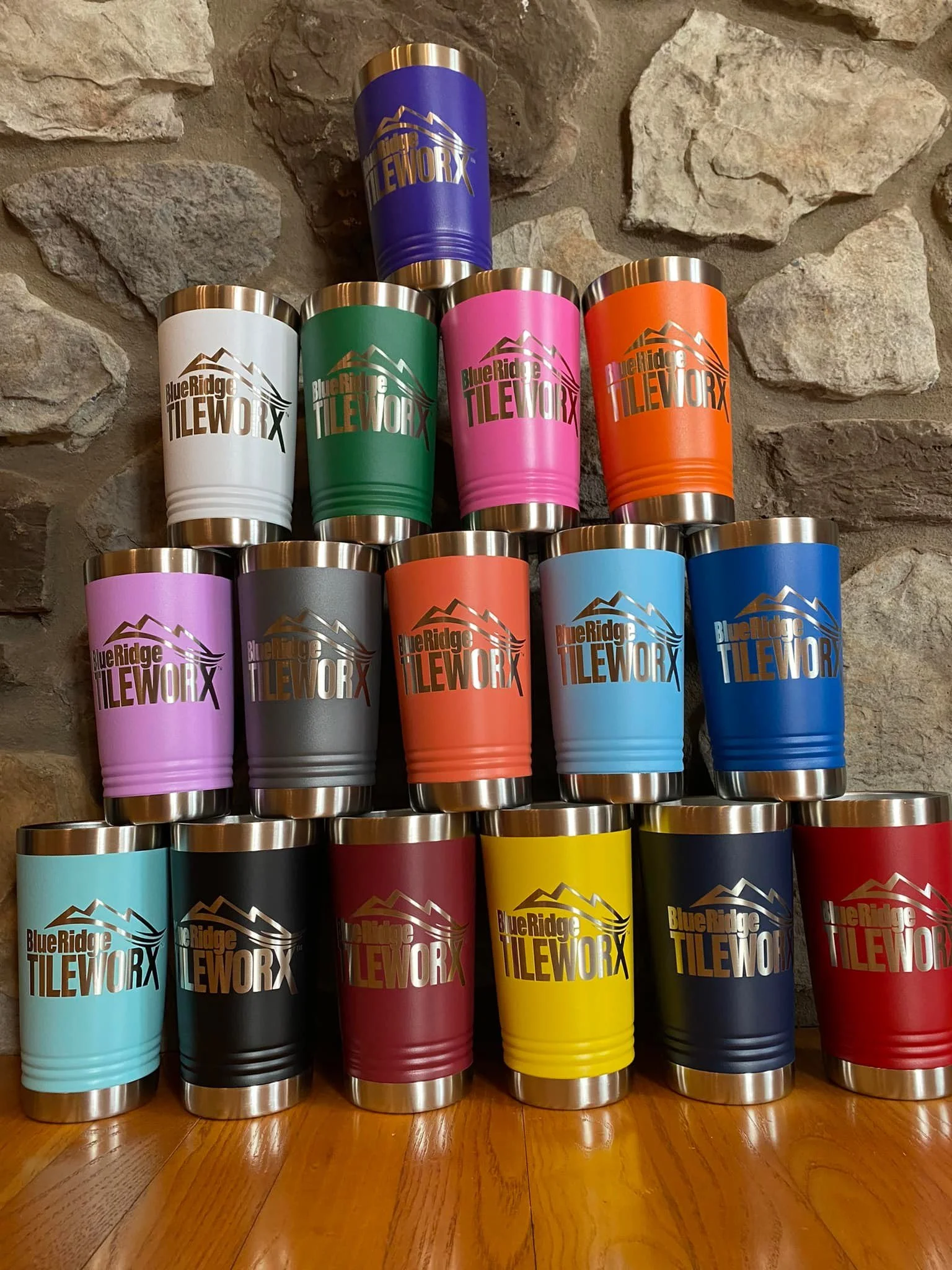 A pyramid of colorful insulated travel mugs with the logo "Blue Ridge TILEWORX" on a wooden floor against a stone wall.