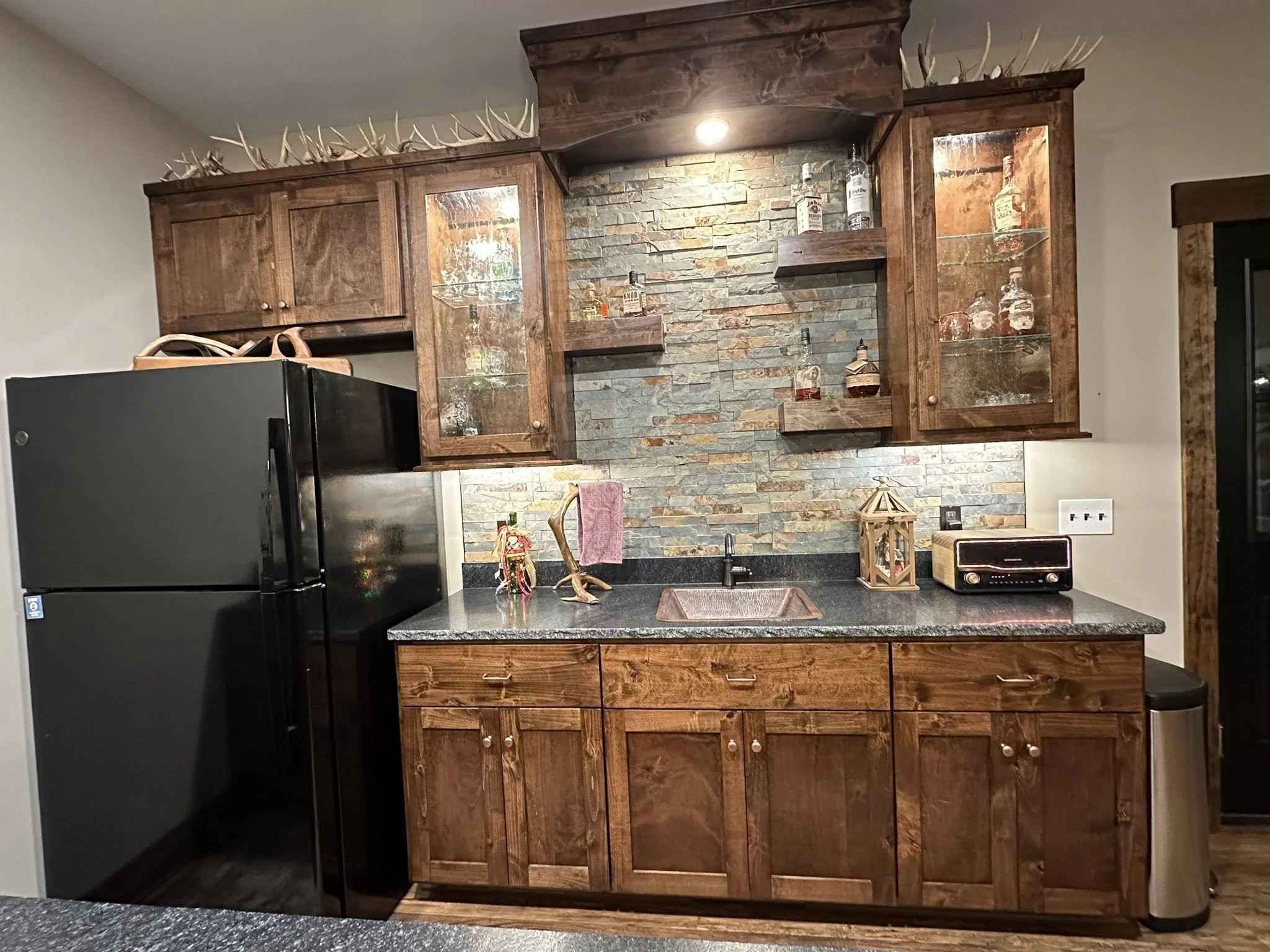 Rustic kitchen with wooden cabinets, black refrigerator, stone backsplash, and decorative bottles on glass shelves.