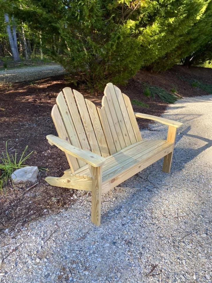 A wooden adirondack-style bench made of unfinished wood, placed on a gravel path near a large green bush with dark mulch underneath.