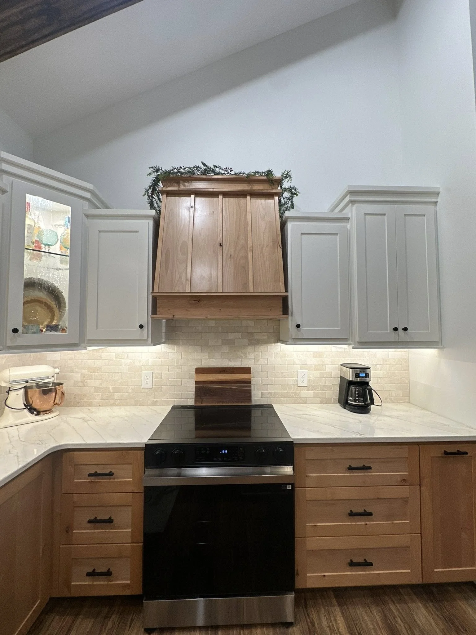 A kitchen with a black electric stove, wooden lower cabinets, white upper cabinets, a white marble countertop, a Coffee Maker, and a stand mixer. There is a wooden range hood with a green garland on top.