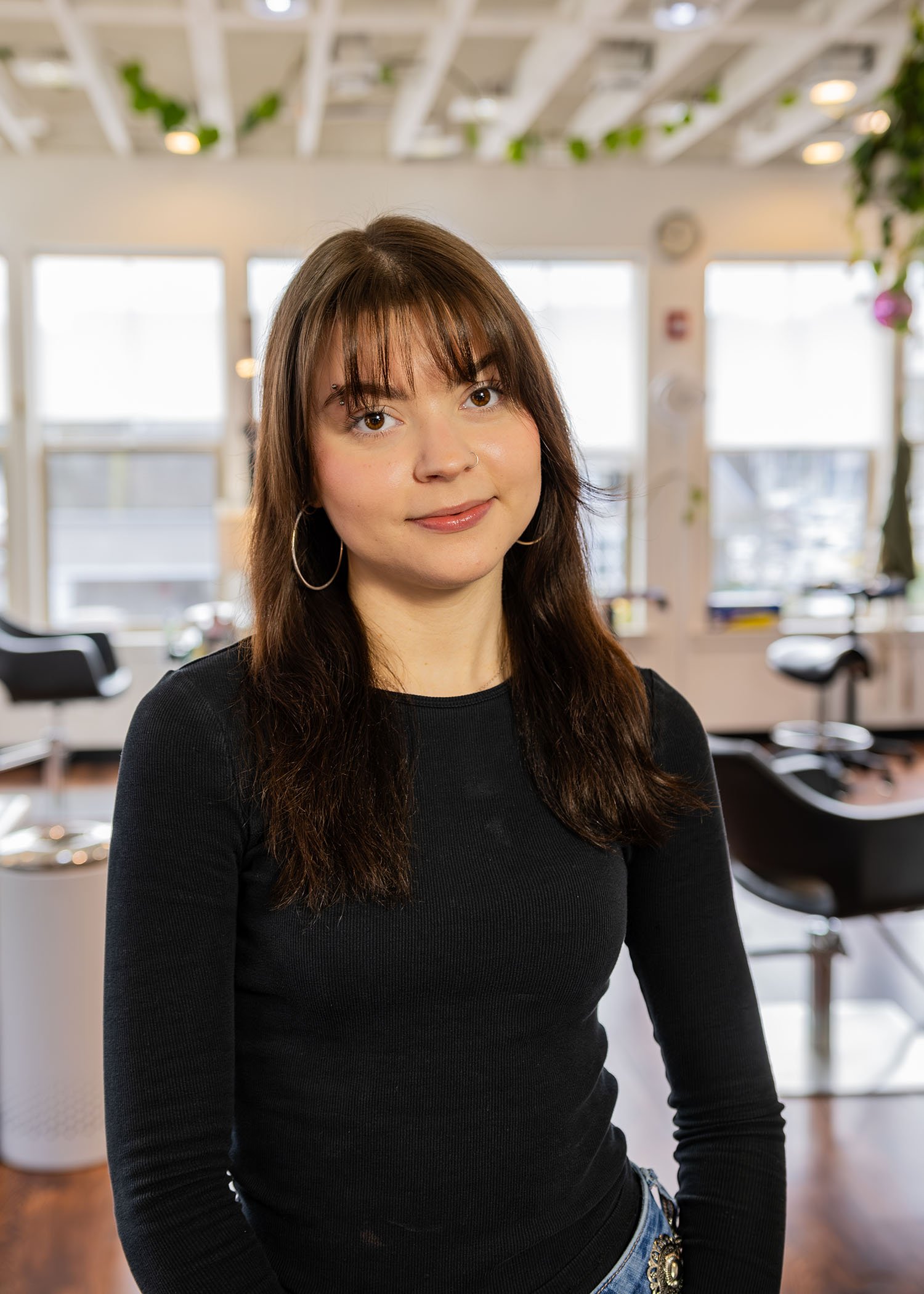 Hair apprentice Jordan with brown hair and hoop earrings smiling in a bright, modern indoor space with large windows and plants.