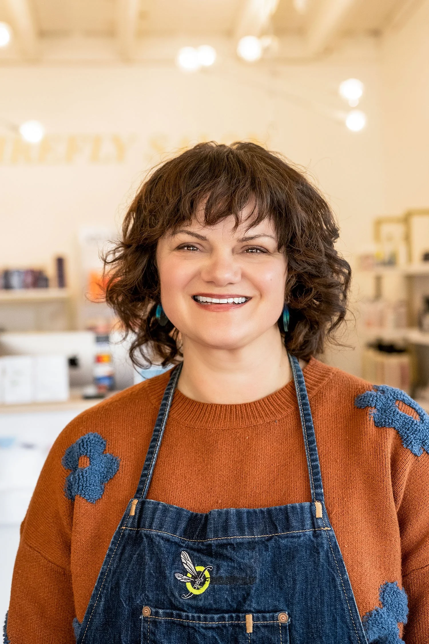 A smiling photo of hair stylist Bronwyn with short curly brown hair wearing a rust-colored sweater with blue decorative patches and a denim apron.