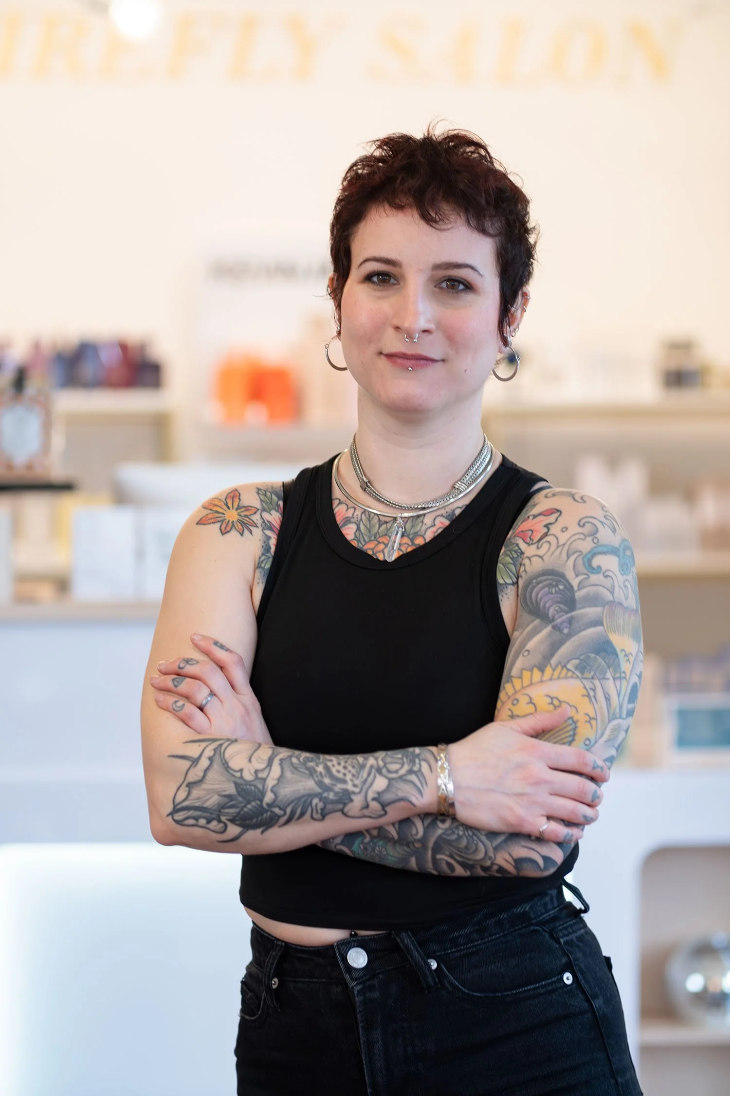 Hair stylist Ashley with short, dark hair and tattoos on her arms and chest is standing with her arms crossed in a retail store or boutique, wearing a sleeveless black top.