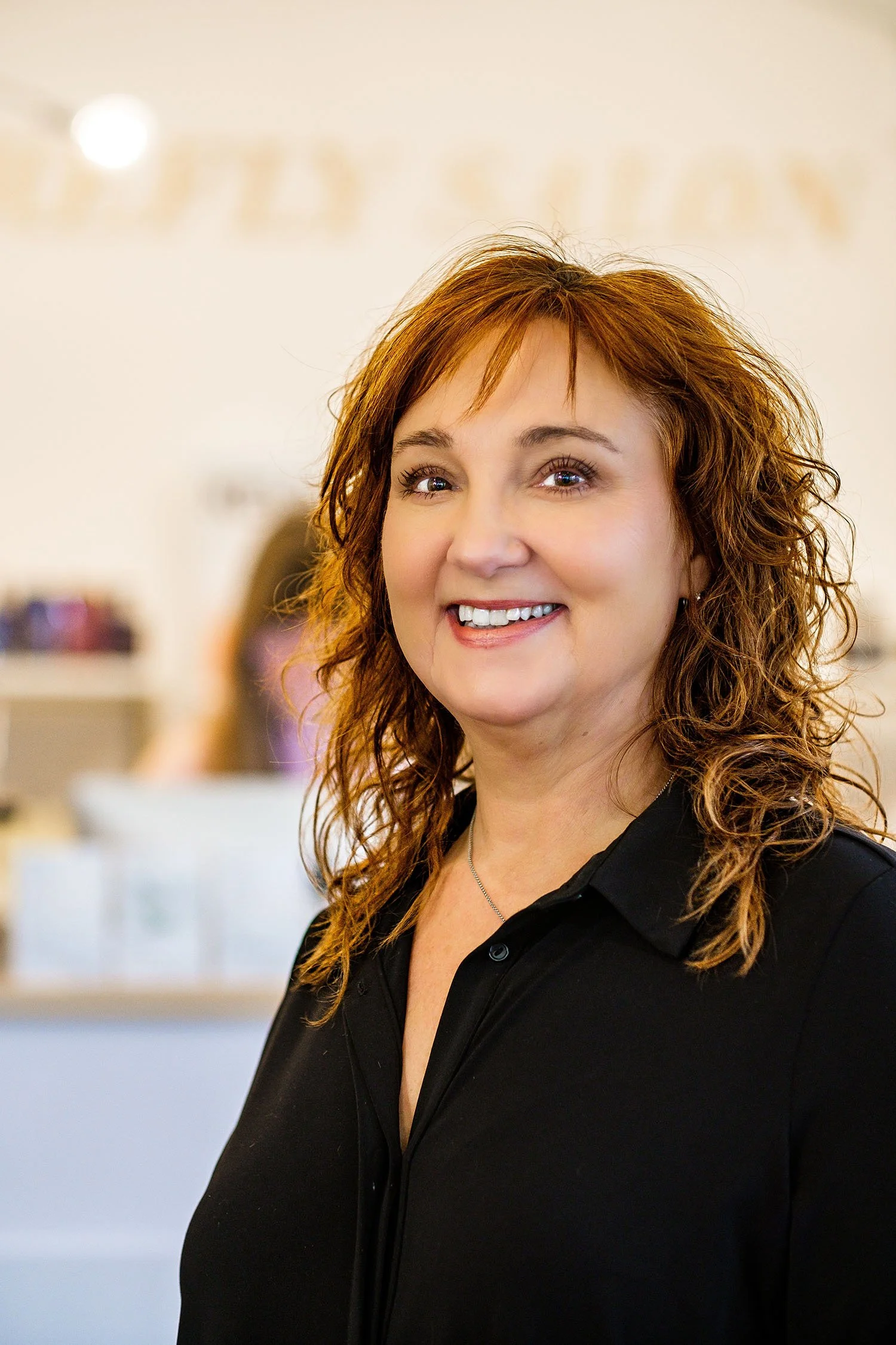 Hair stylist Michelle with curly red hair smiling in an indoor setting, wearing a black shirt.