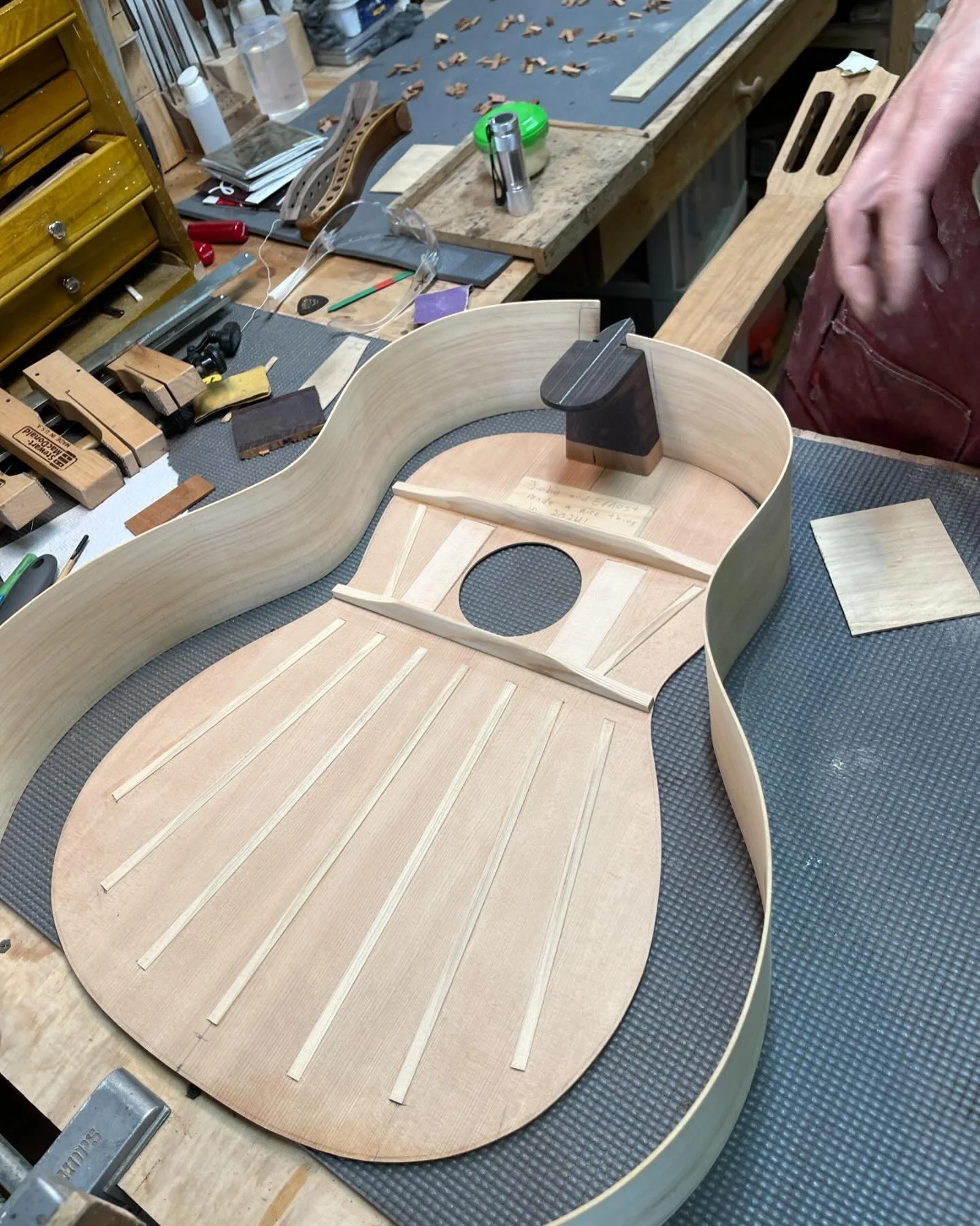 Close-up view of a guitar body in the process of being built, with the wood frame and internal bracing visible, on a workbench among various woodworking tools and materials.