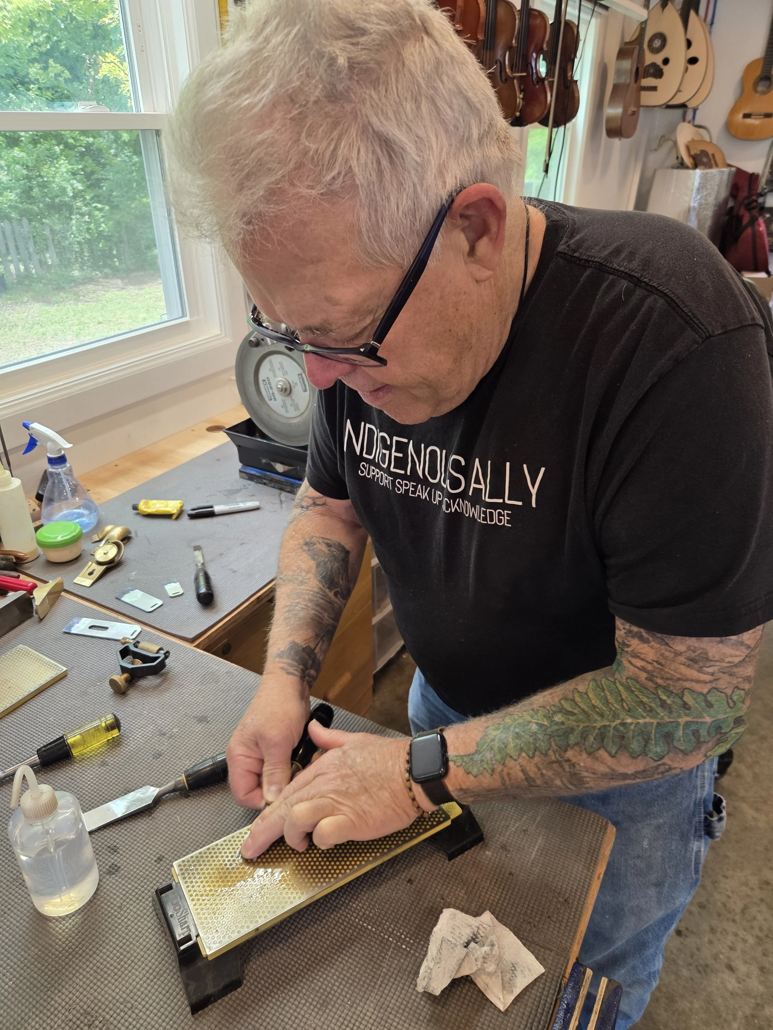 A man with tattoos and glasses working on a soldering project at a workbench with various tools, a knife, and a soldering iron, in a room with multiple guitars hanging on the wall and a window showing greenery outside.