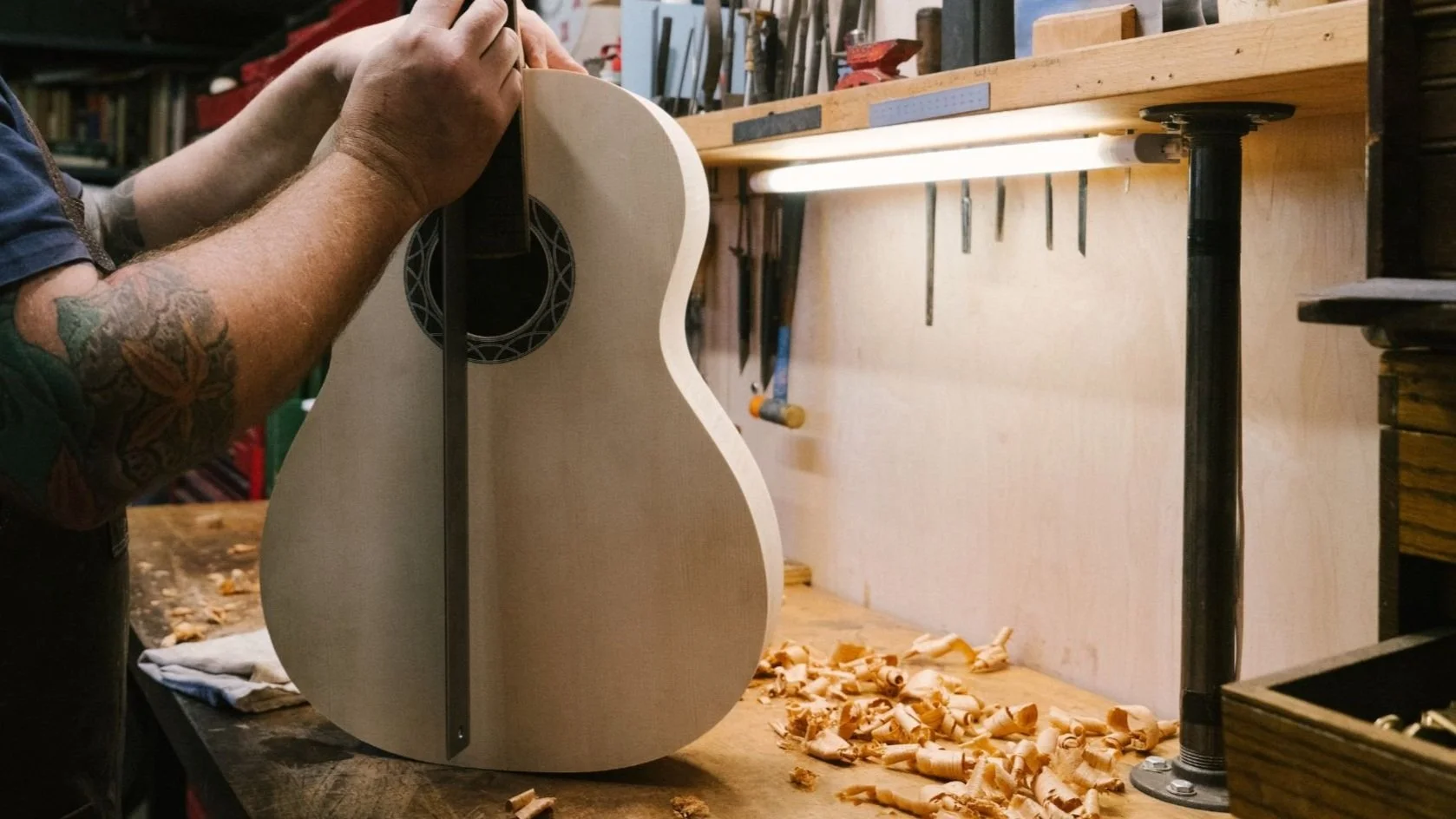 A person working on building an acoustic guitar in a woodworking shop, with wood shavings on the workbench.