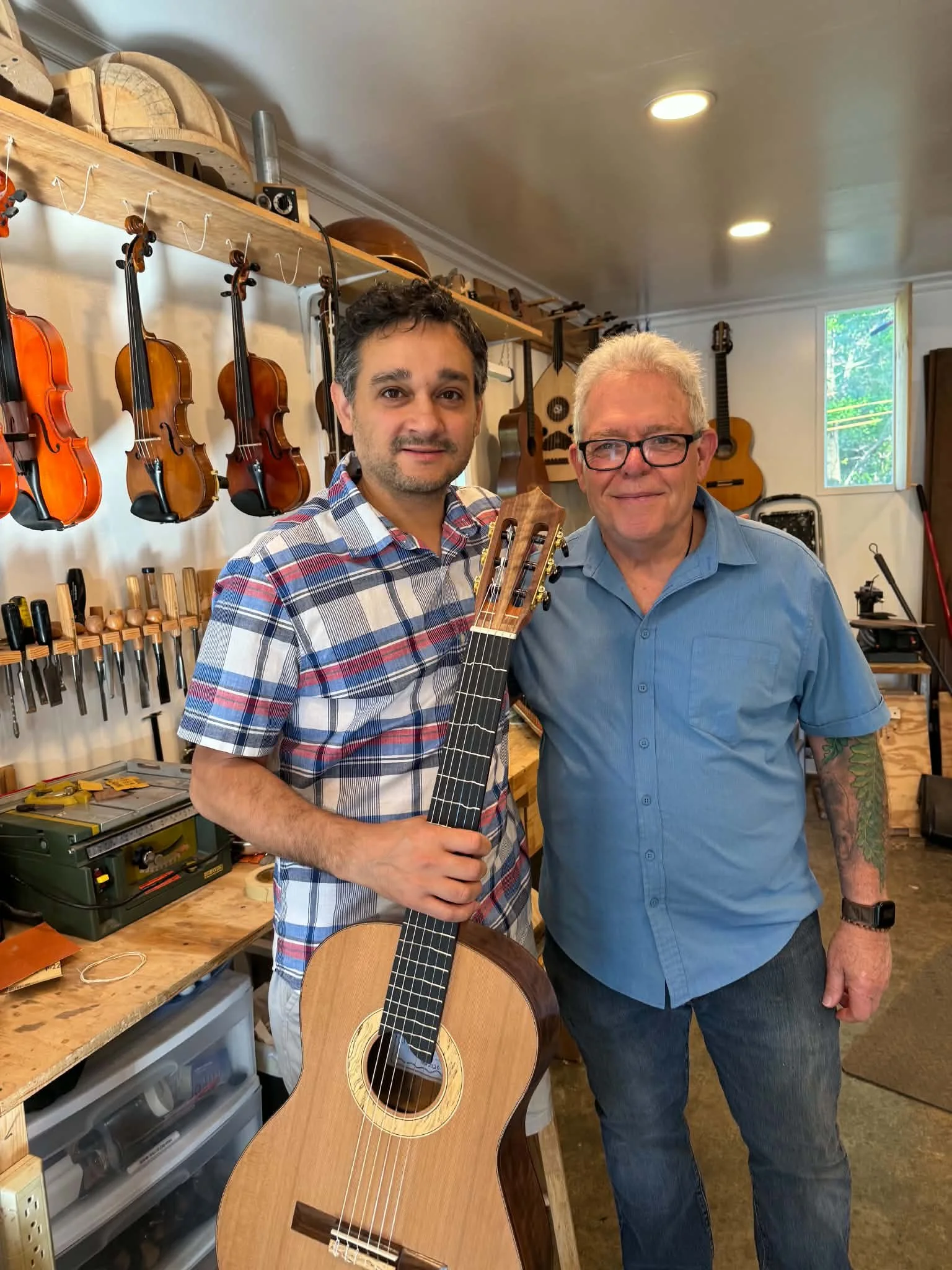 Two men standing in a music workshop with violins hanging on the wall behind them. One man is holding an acoustic guitar, and both are smiling and looking at the camera.