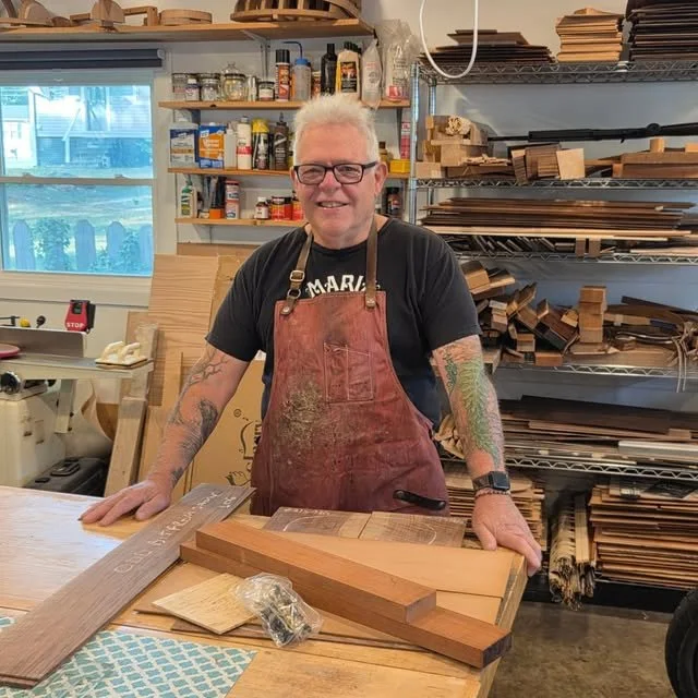 Smiling man with white hair, glasses, tattoos on arms, wearing a black T-shirt and a red apron, standing in a woodworking shop with wood pieces and tools in the background.