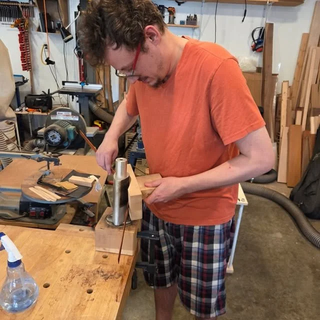 A young man with glasses working on a woodworking project in a workshop, using a clamp on a piece of wood.