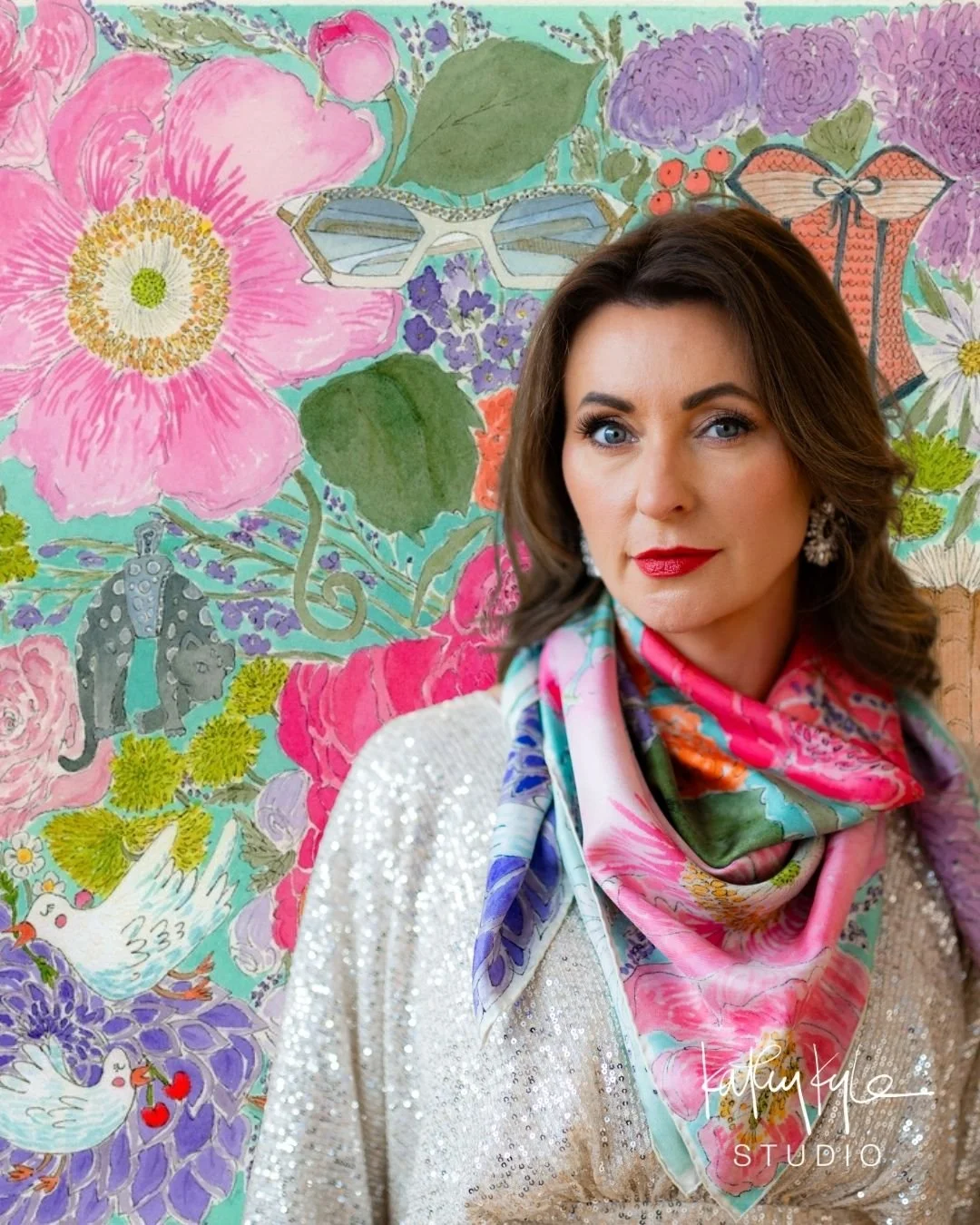 A woman with dark hair, red lipstick, and blue eyes wearing a shimmery white top and a colorful floral scarf stands in front of a vibrant, botanical-themed background mural featuring large pink, purple, and white flowers, green leaves, and illustrations of animals and insects.