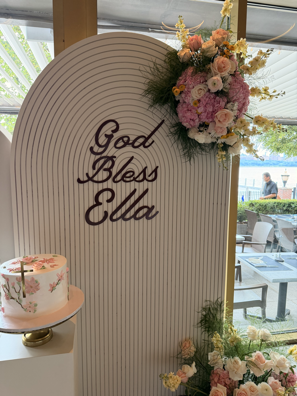 Decorative display with pink and white flowers, a cake decorated with pink flowers, and a sign that reads 'God Bless Ella' at a celebration event.