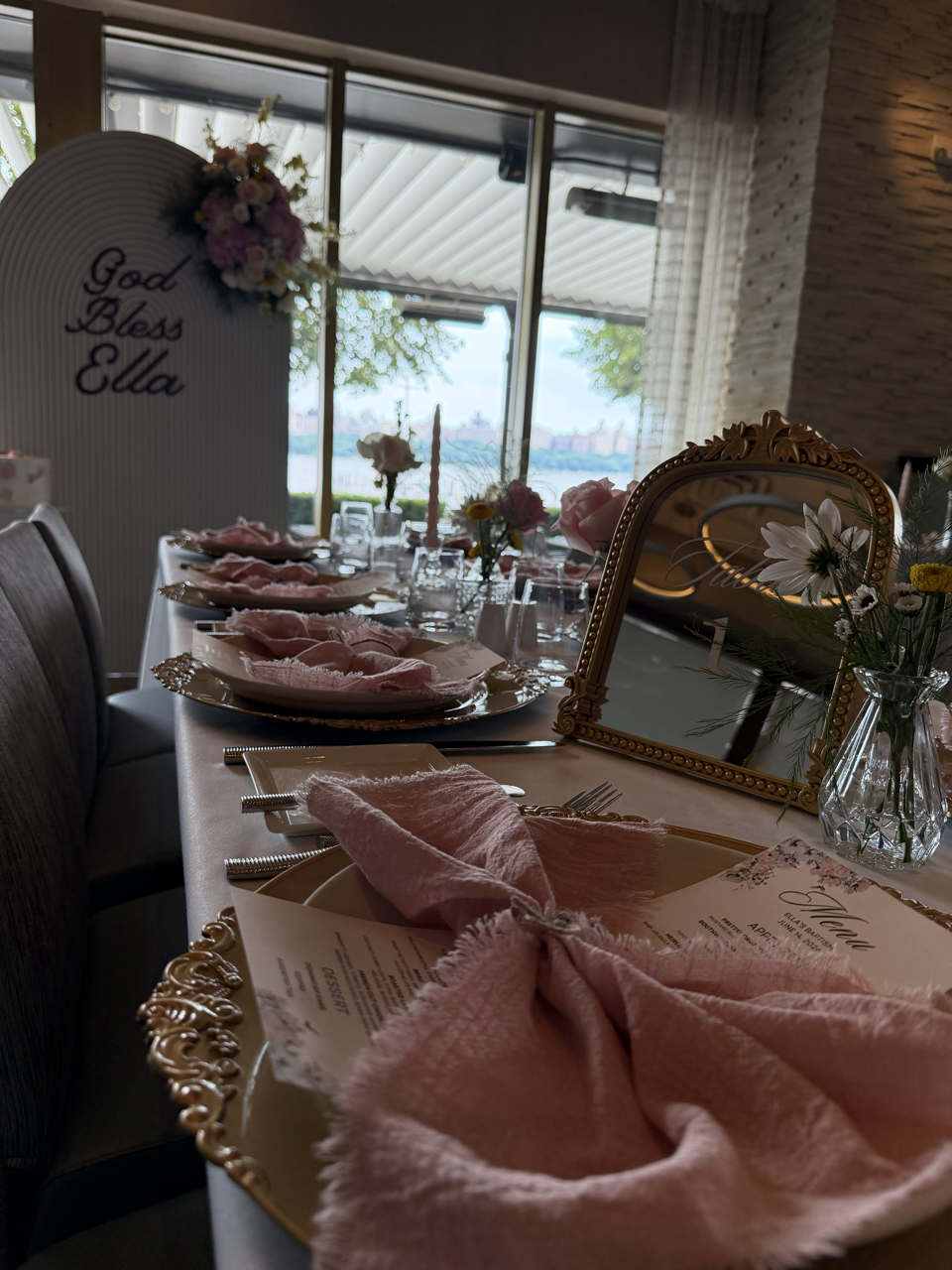 Elegant dining table set for a celebration with pink napkins, floral arrangements, a mirror, and a display board reading 'God Bless Ella,' near a large window overlooking a river or body of water.