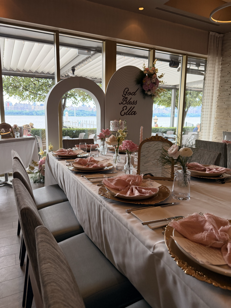Decorated dining table with pink napkins, flowers, and a wine glass, set for a celebration at a restaurant with a view of a river outside.