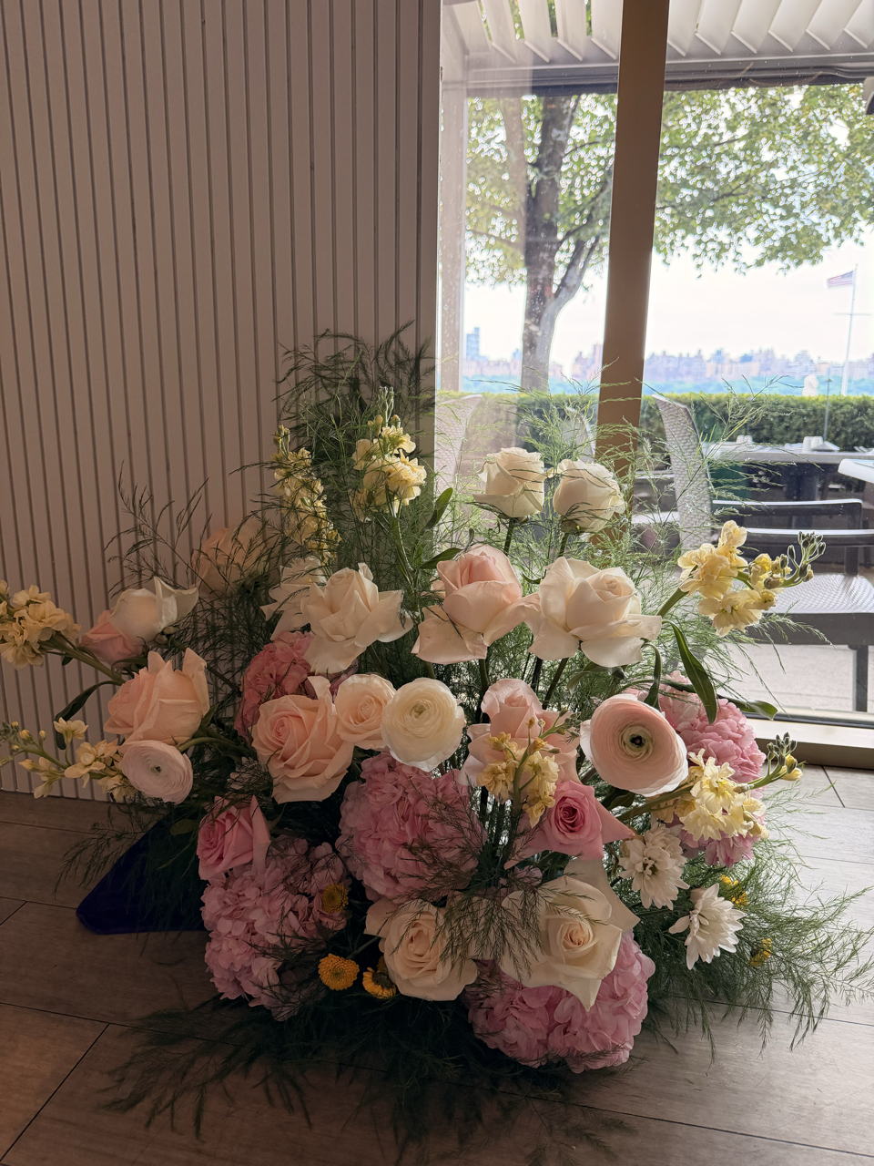 A large arrangement of pink, white, and yellow flowers sitting on a wooden floor near a window with outdoor tables and chairs visible.
