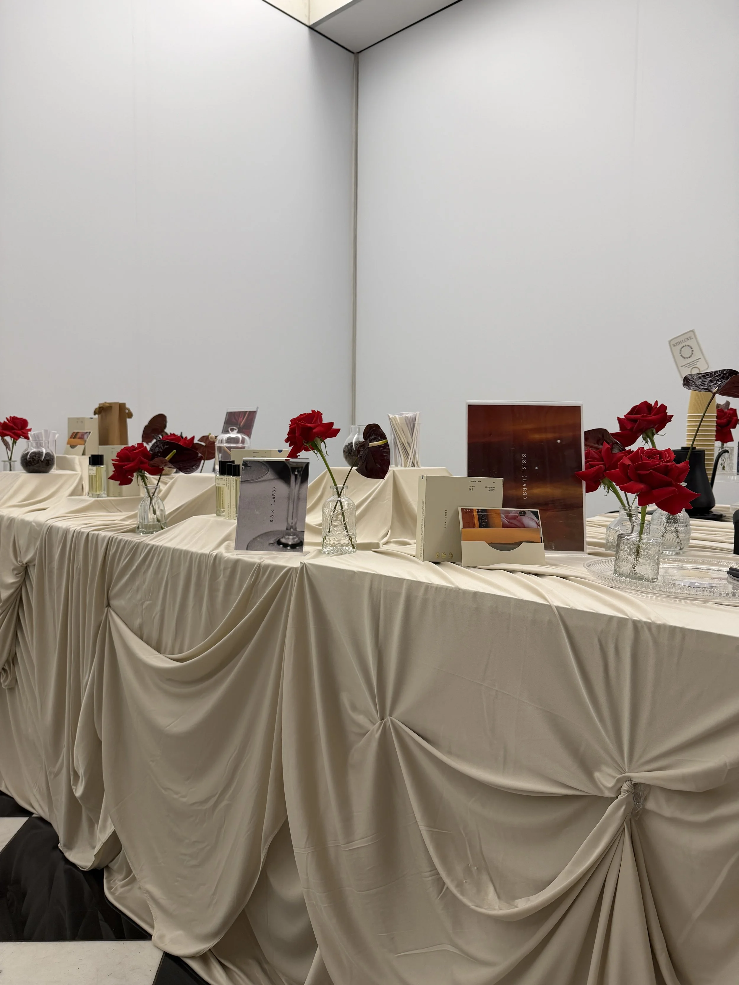 A table draped with beige fabric, decorated with red roses in glass vases, and various books and decorative items displayed on it, in a plain white room.