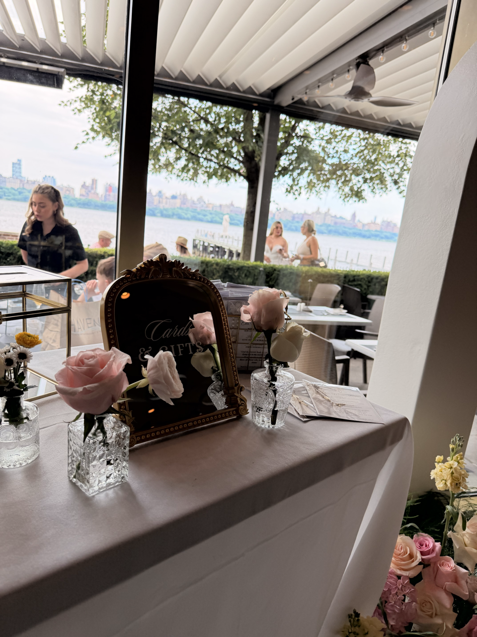 Decorated table with pink and white roses in glass vases, a small mirror, and arrangements for a celebration, with a view of the river and people outside near a tree.
