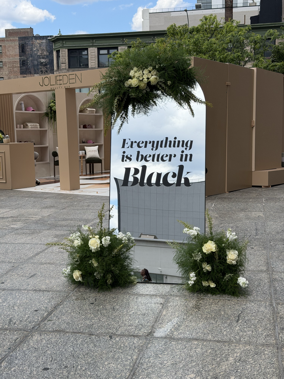 A mirror sign with a floral arrangement on top and at the base, reflecting a building and sky with clouds, displays the text "Everything is better in Black" on a city sidewalk area