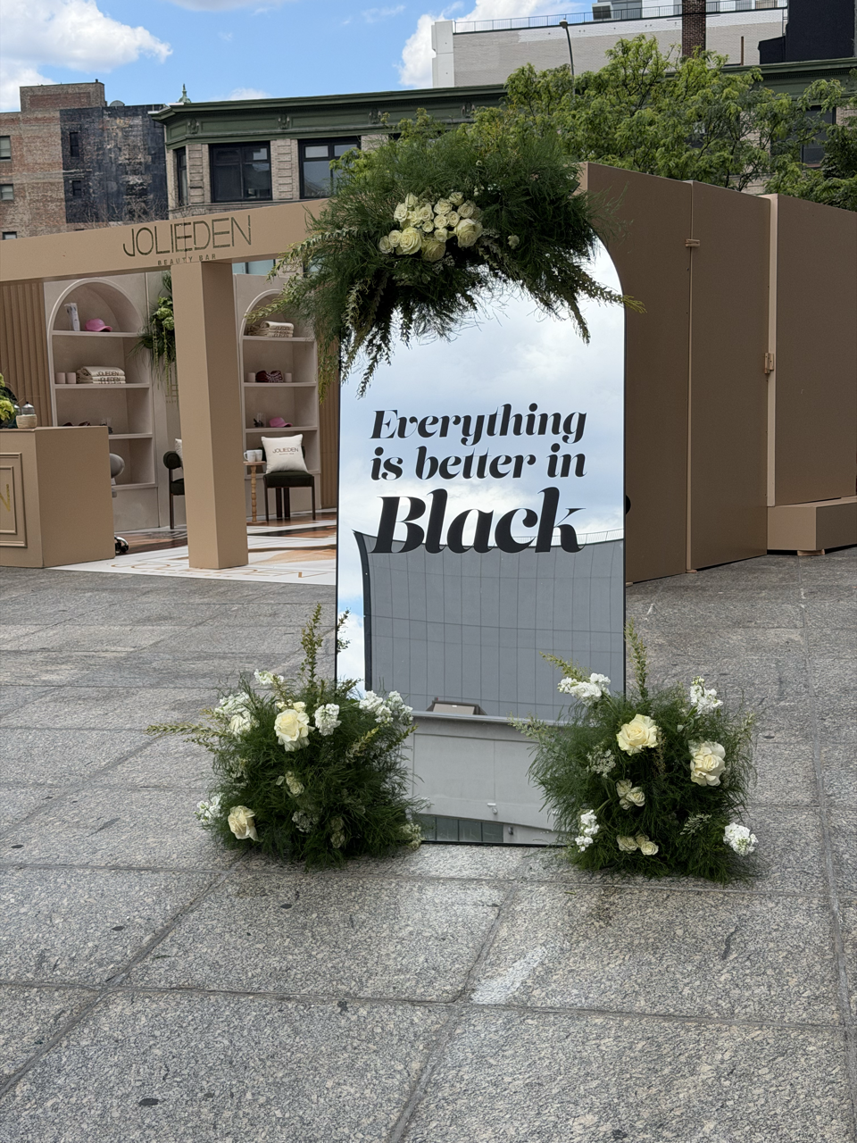 A sign with the message 'Everything is better in Black' surrounded by white flowers and greenery, with a backdrop of a storefront and city buildings.