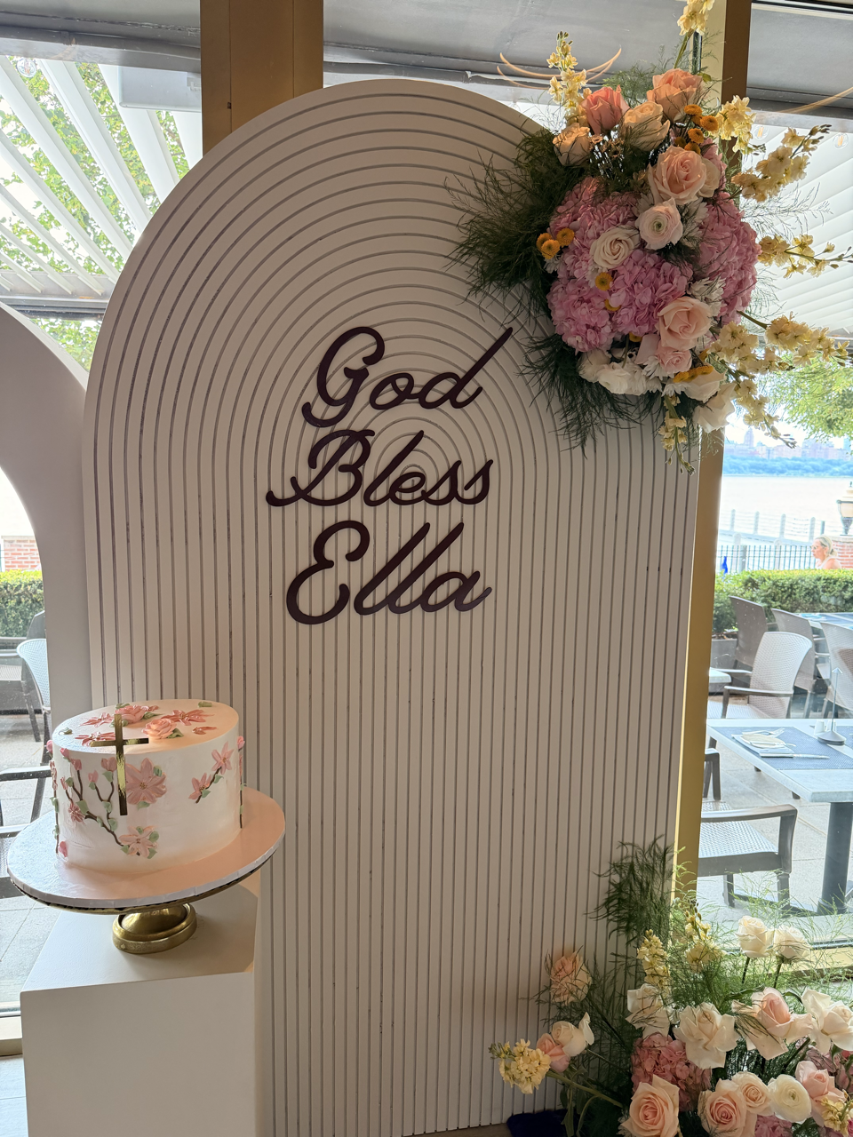 A decorated display with a floral arrangement and a cake, featuring a white panel with the phrase 'God Bless Ella' written in cursive, surrounded by pink and white flowers with greenery in a bright room near a window.