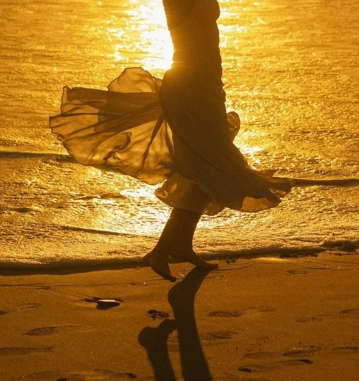 A woman in a dress dancing on the beach at sunset, with her silhouette reflected in the wet sand.