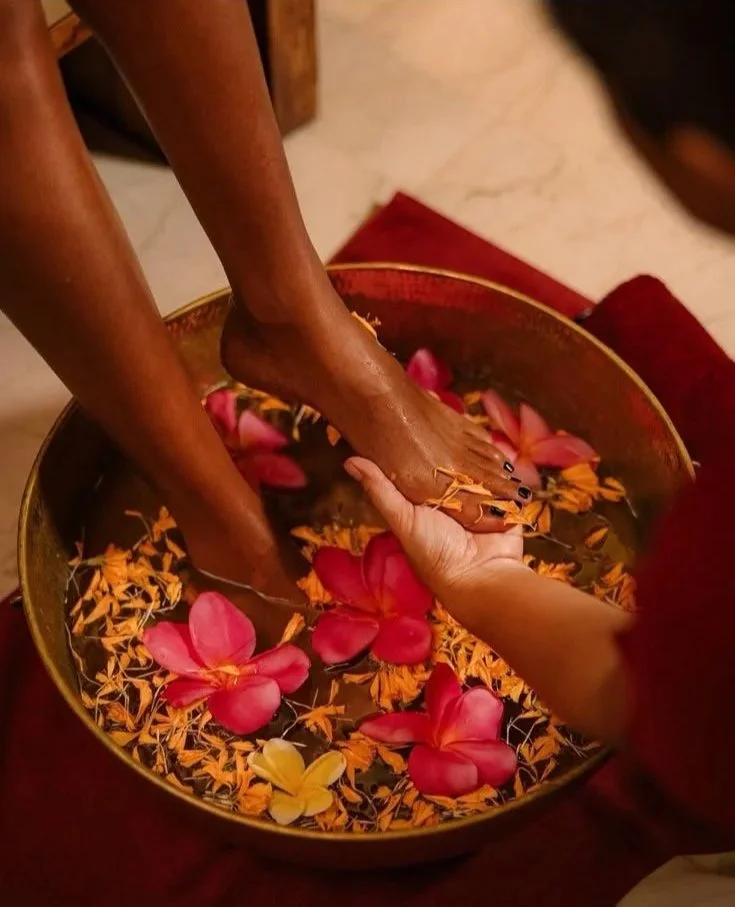 Person receiving a foot bath in a bowl filled with flower petals and water while sitting on a red cloth.