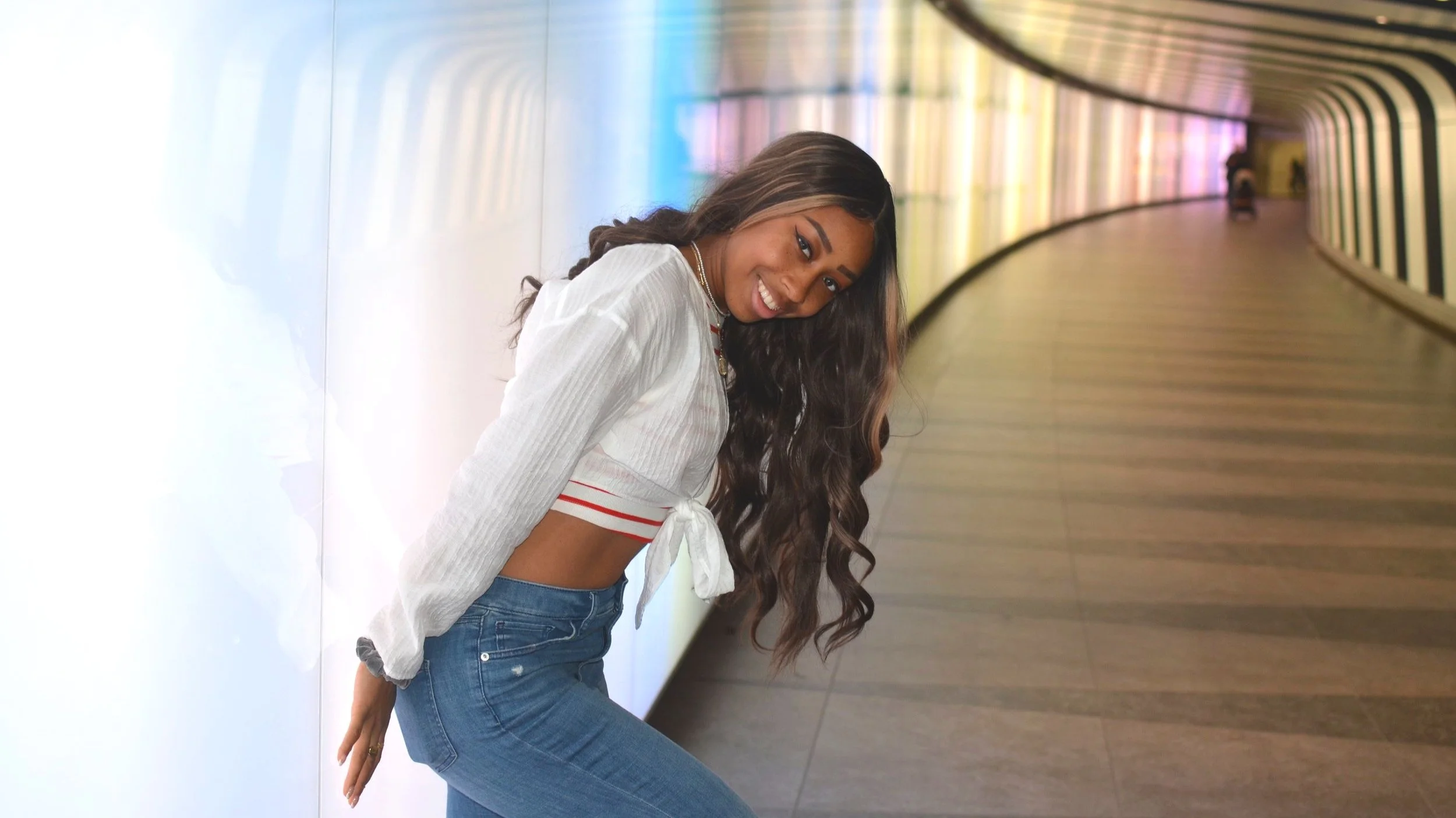 A young woman with long wavy hair smiling, wearing a white cropped top with red accents and blue jeans, sitting against a colorful, illuminated corridor wall.
